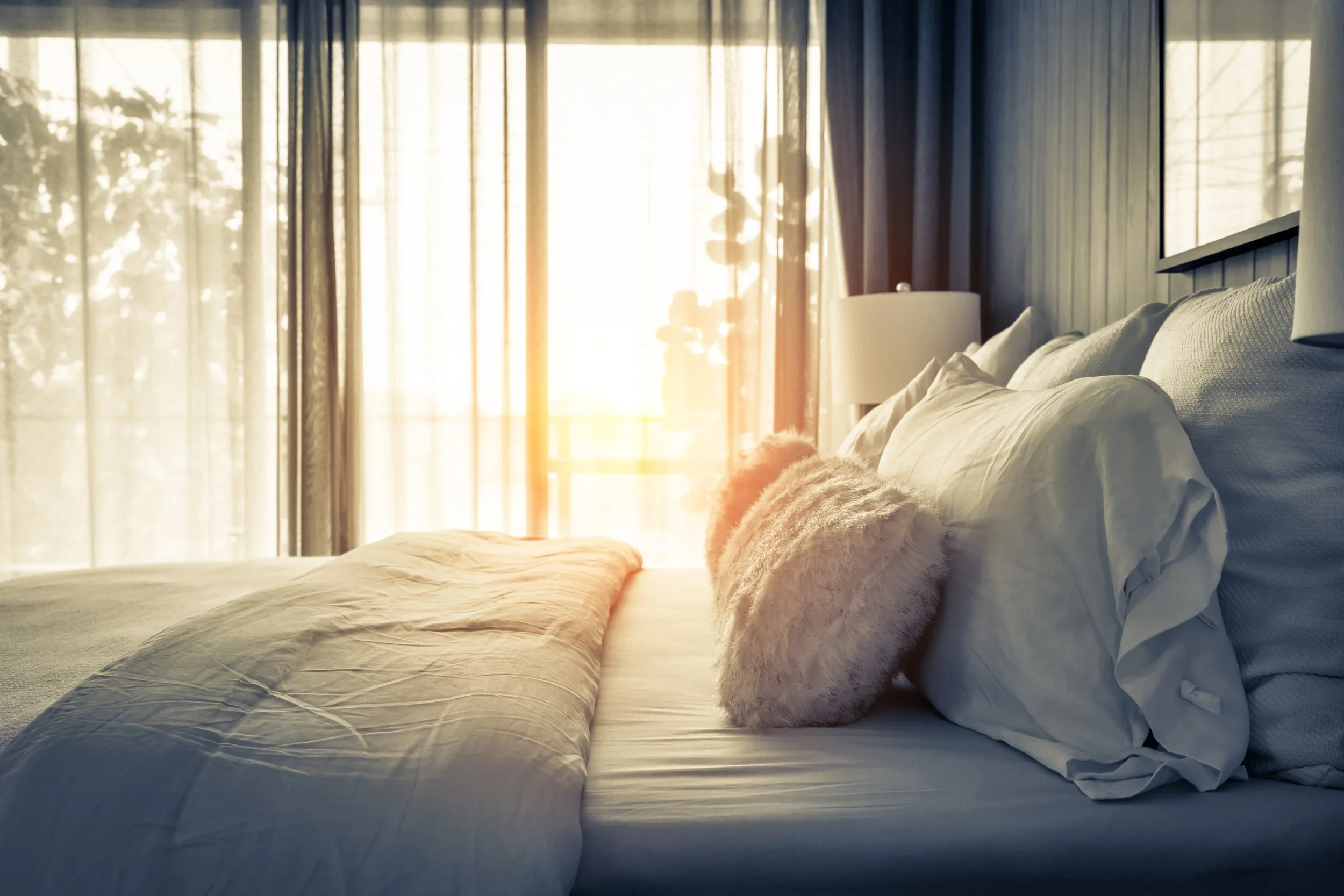 A neatly made bed with white pillows and a fluffy round cushion, illuminated by sunlight coming through large glass doors with curtains, in a bedroom.