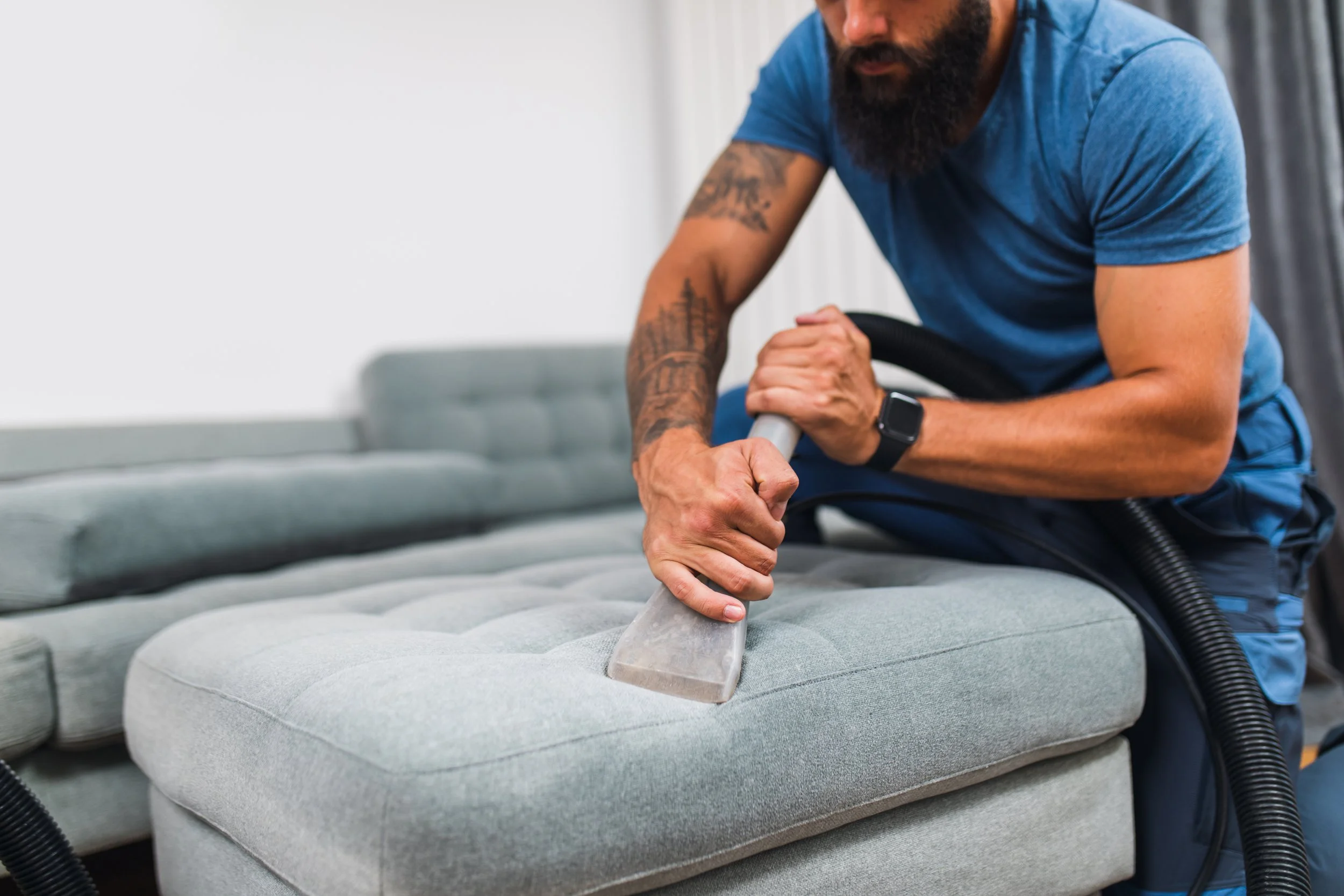 A man with a beard and tattoos cleaning a light gray upholstered couch with a handheld vacuum cleaner.