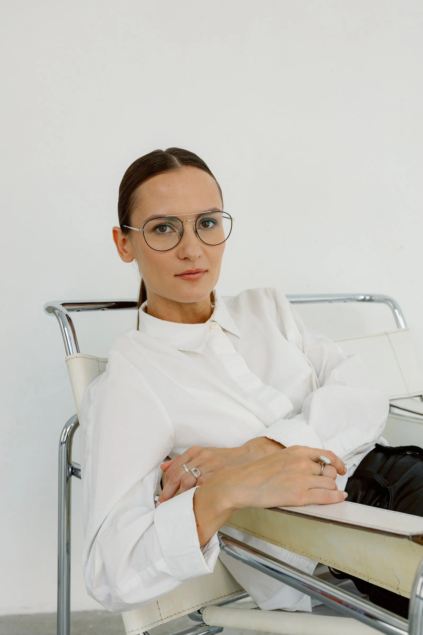 A woman with brown hair pulled back, wearing glasses and a white shirt, sitting in a chair with her arms crossed on a table, against a plain white wall.