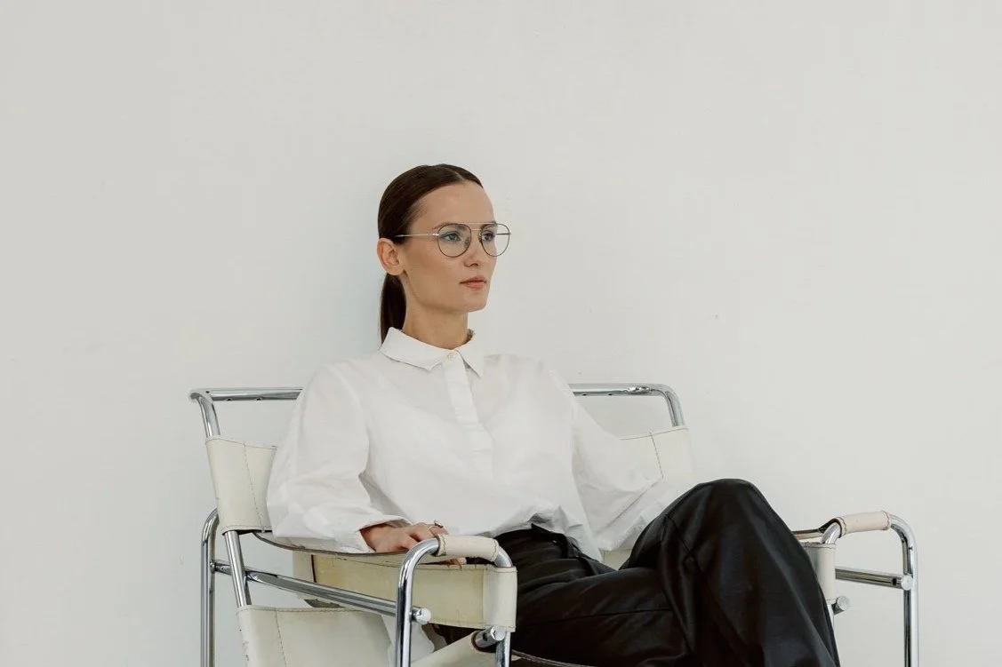 Woman with glasses sitting in a wheelchair against a plain white wall.