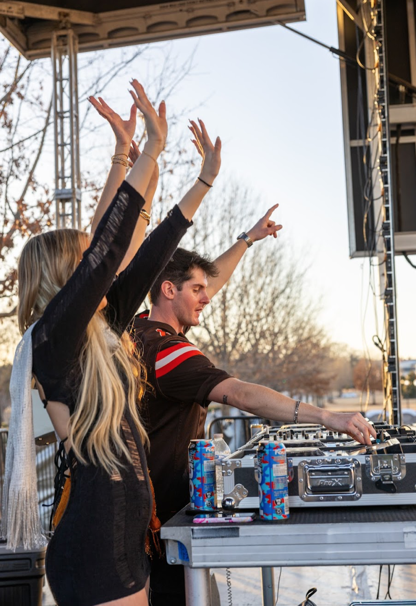 DJ performing outdoors with a woman dancing beside the mixing console, both raising their hands in the air, with drinks on the table.