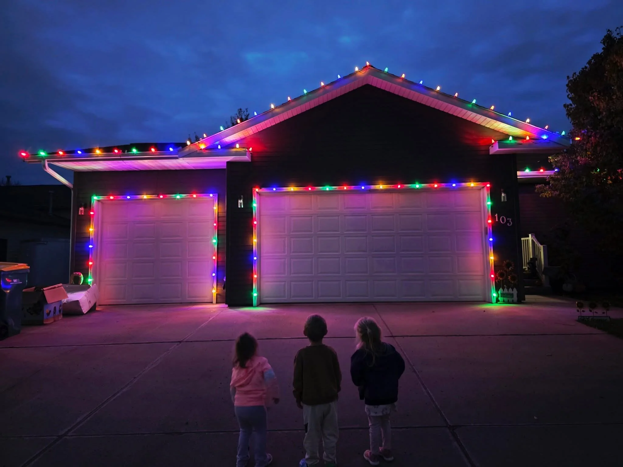 Three children standing in front of a house decorated with multicolor C9 LED Christmas lights – professional holiday lighting in Mandan