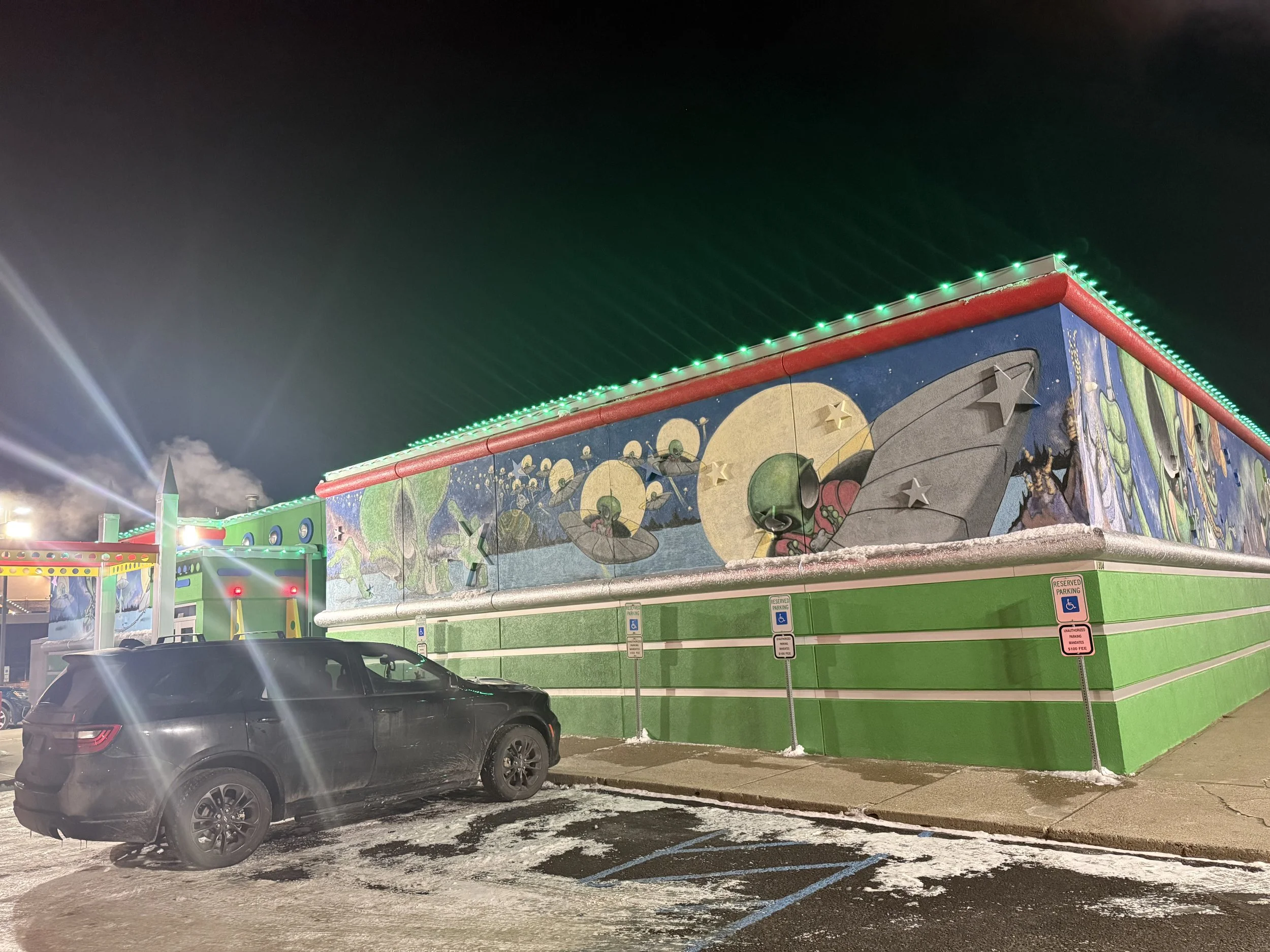 Nighttime scene of a colorful, decorated building with space-themed murals, green and red trim, and green string lights, with a black SUV parked in a snowy lot with reserved parking signs.