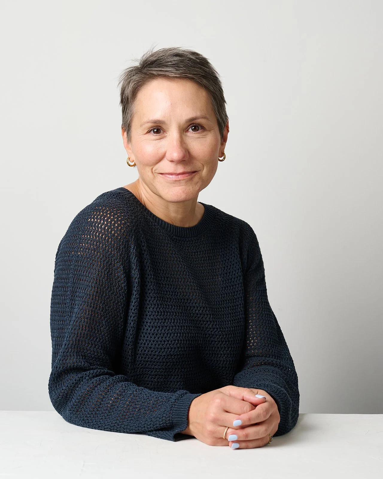A woman with short hair, wearing a black sweater, sitting at a white table, smiling, against a plain light gray background.