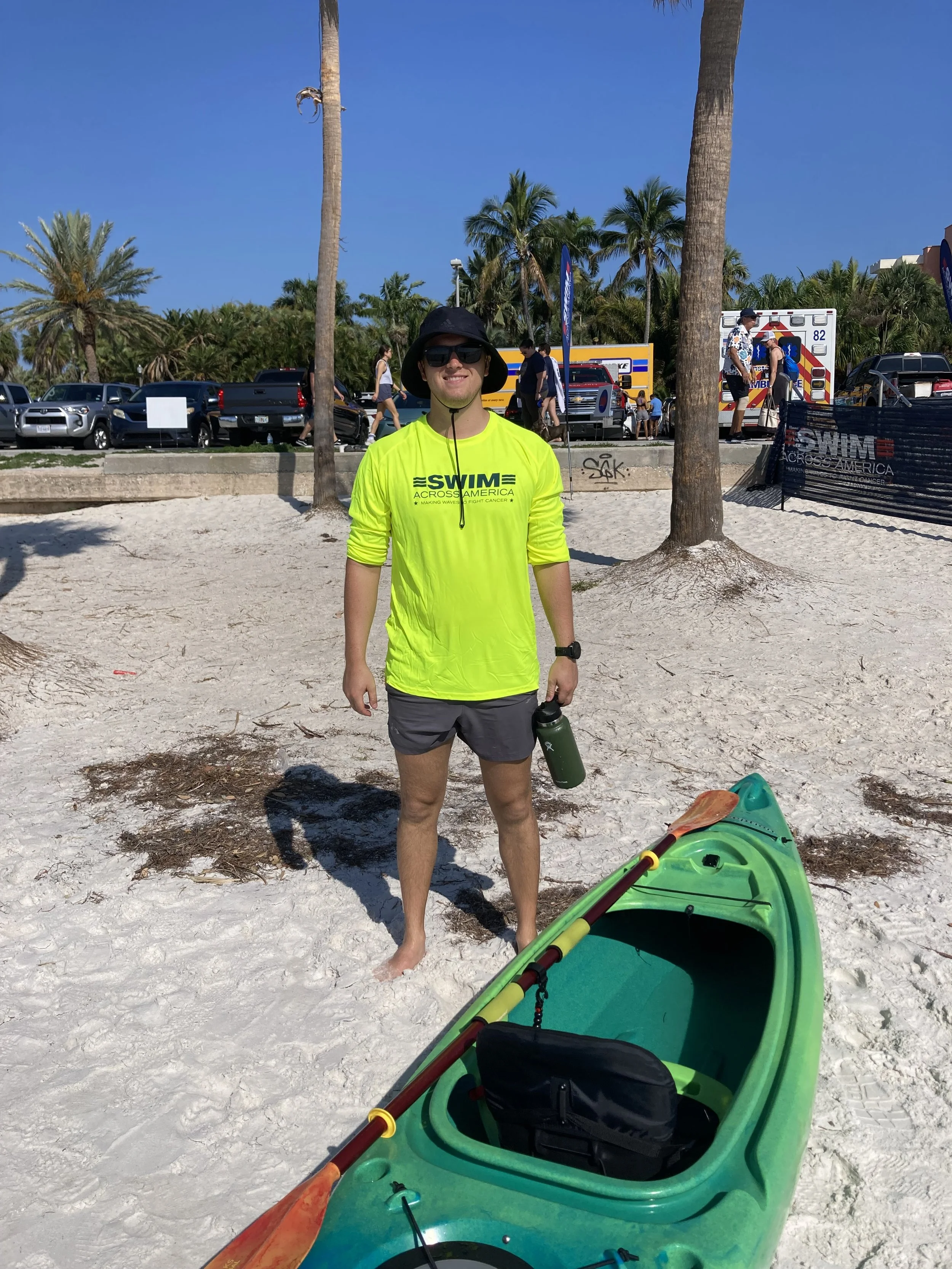 A man wearing a bright yellow swim shirt, black hat, and sunglasses stands on a sandy beach with a green kayak and paddle in the foreground. In the background, there are palm trees, parked cars, an ambulance, and several people walking and standing.