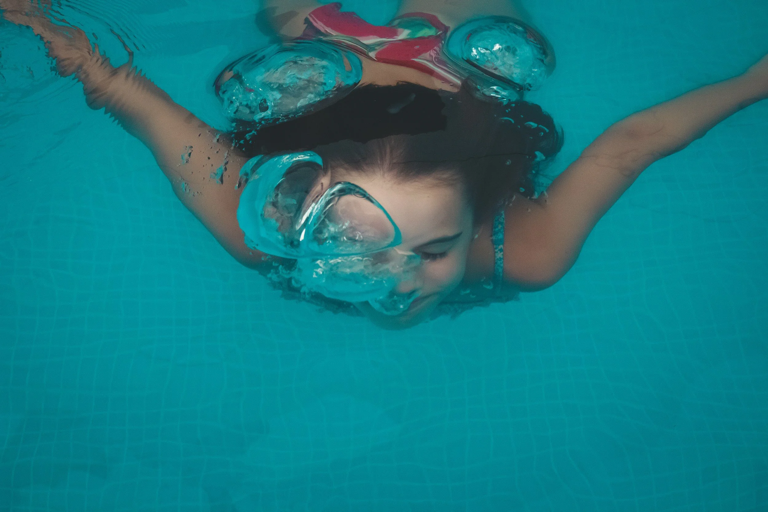A person swimming underwater in a pool with goggles and a swimsuit, with arms extended forward and water bubbles around.