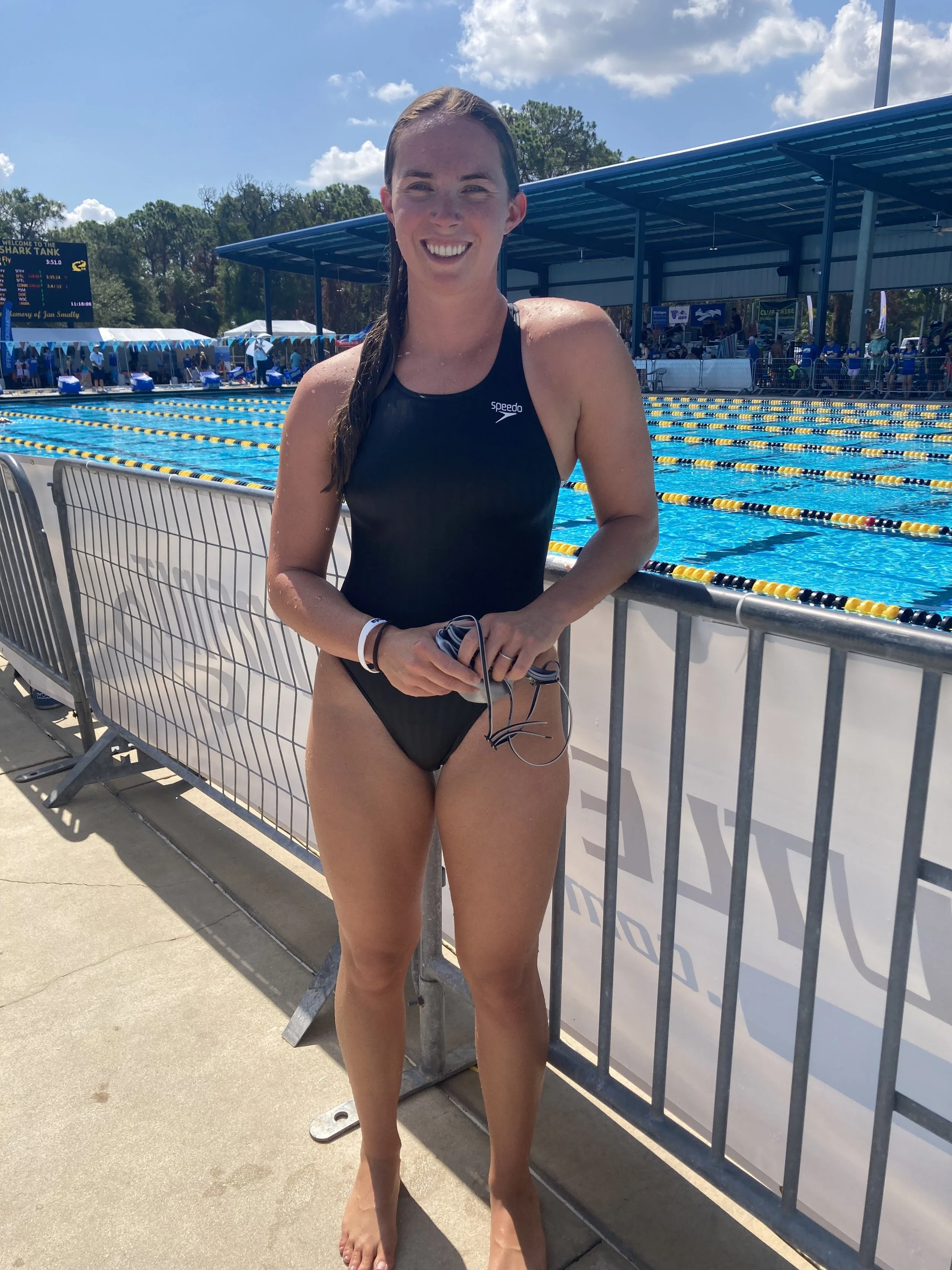 A woman in a black swimsuit standing near a swimming pool with a big smile, holding goggles. Behind her is a pool with yellow and black lane dividers, a scoreboard, and a covered spectator area under blue sky with clouds.