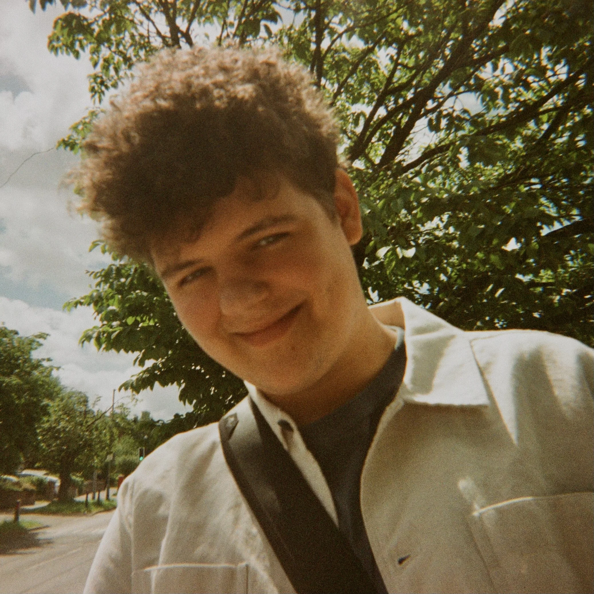 Joshua Bibby, with short curly hair and light skin, wearing a white shirt, is smiling and standing outdoors near a tree. The background shows a cloudy sky and a street scene.