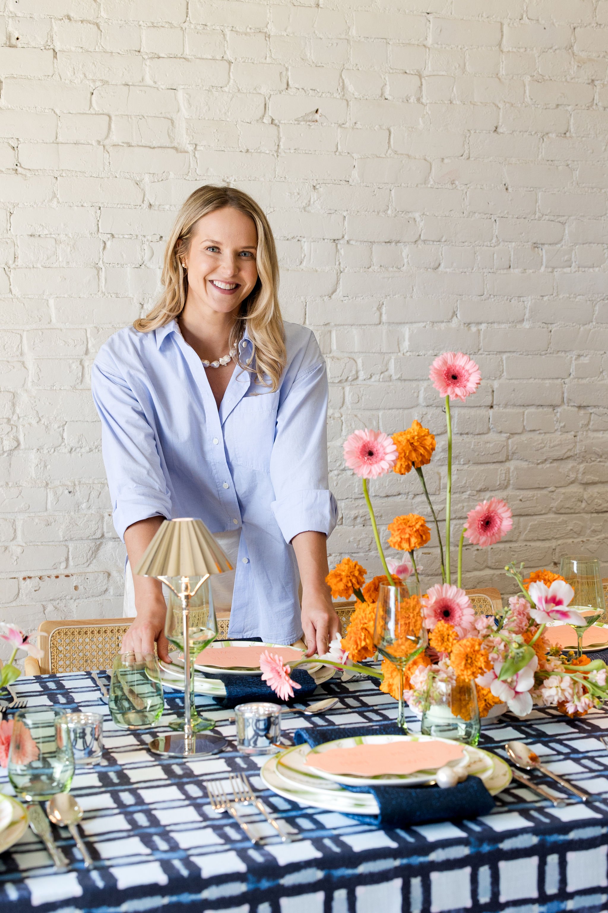 Woman in a light blue shirt setting a table for a meal with colorful flowers as centerpiece.