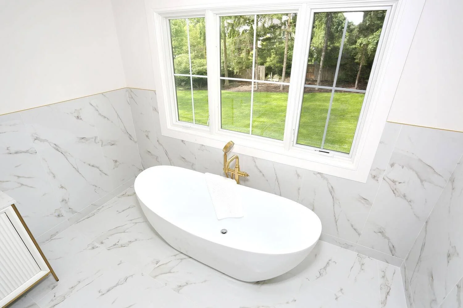 Freestanding bathtub beneath large window with marble tile accents in a bright primary bathroom by Dadak LLC, Park Ridge NJ.