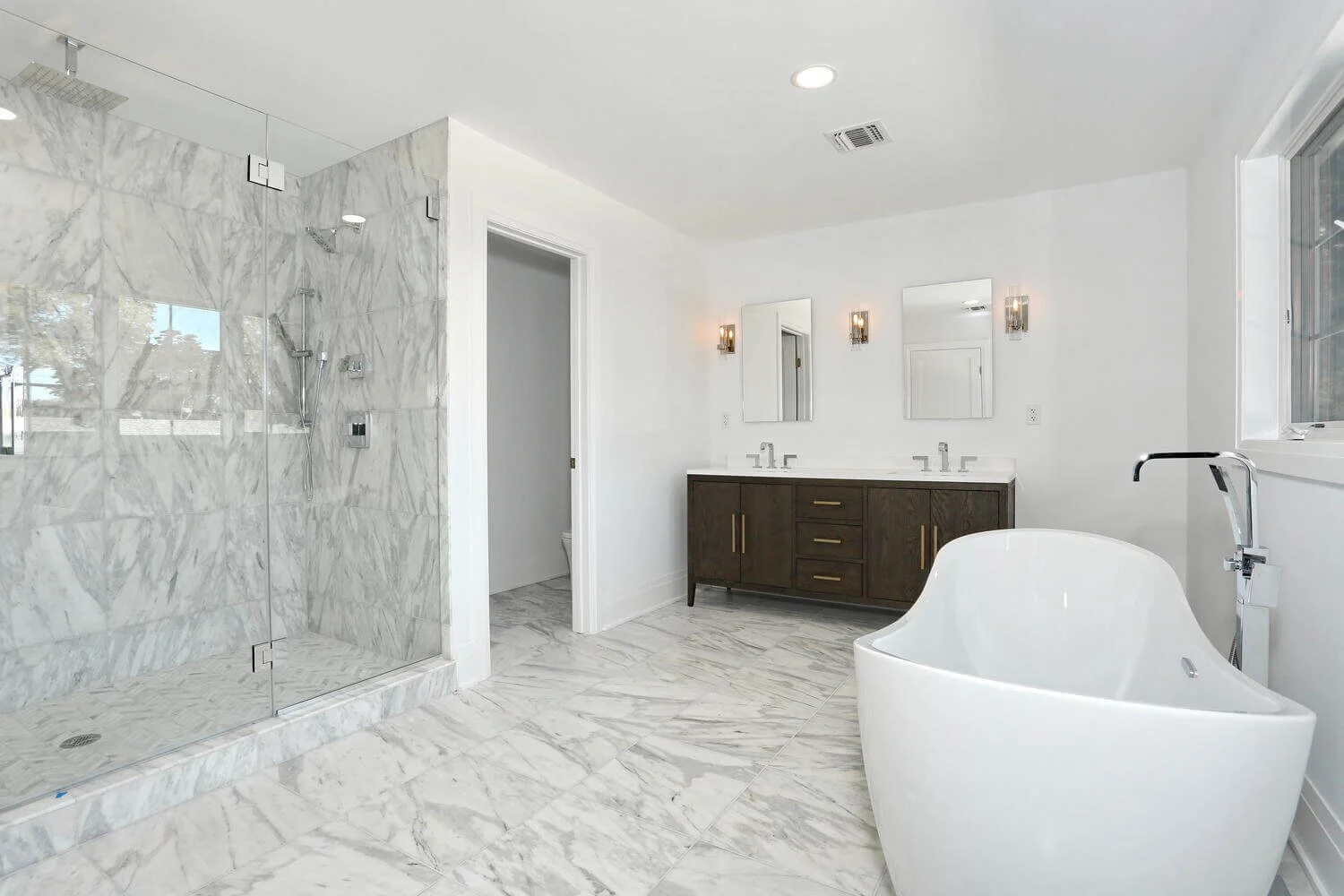 Luxurious primary bathroom with marble tile, glass-enclosed shower, double vanity, and freestanding soaking tub in a custom River Vale home by DADAK LLC.