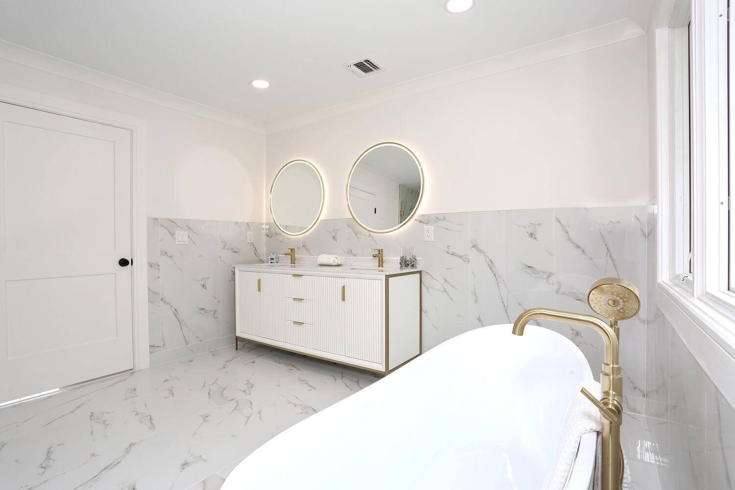 Freestanding tub and illuminated double vanity with marble tile finishes in a Park Ridge modern farmhouse by Dadak LLC.