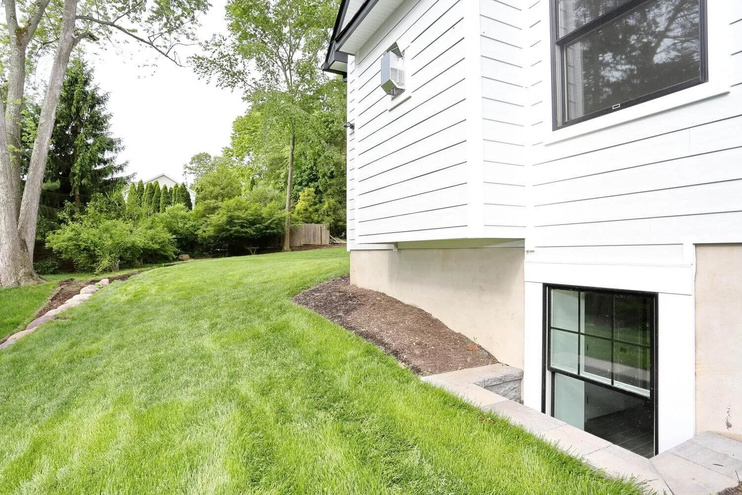 Side exterior of modern farmhouse with basement window, white siding, and landscaped green lawn in Park Ridge by Dadak LLC.
