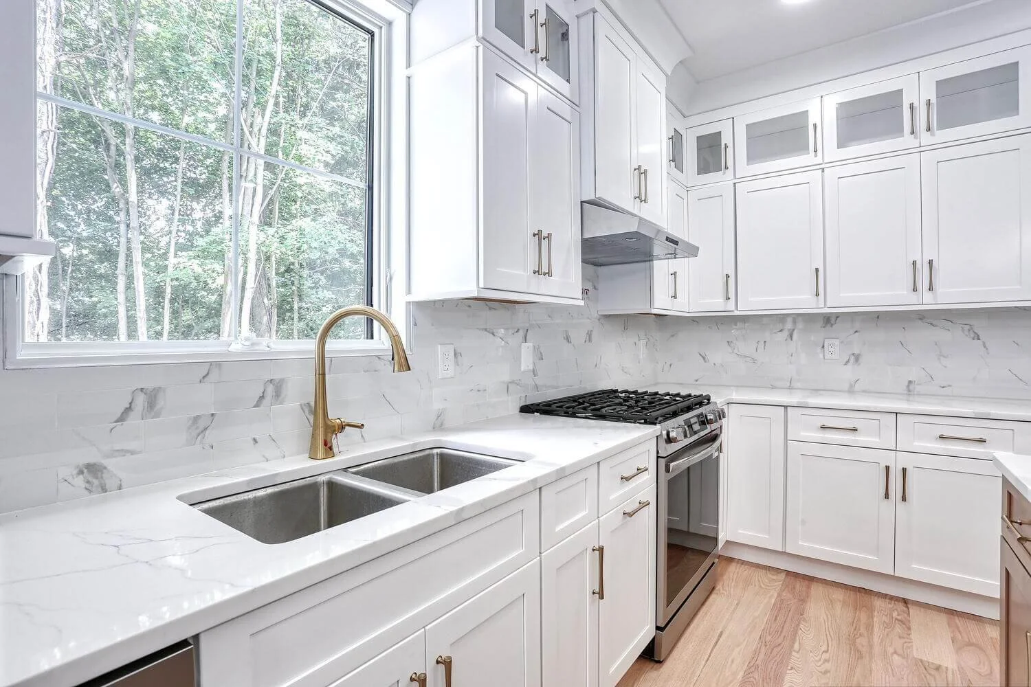 Kitchen sink and gas range with white shaker cabinets, marble tile backsplash, and gold fixtures in Montvale home.