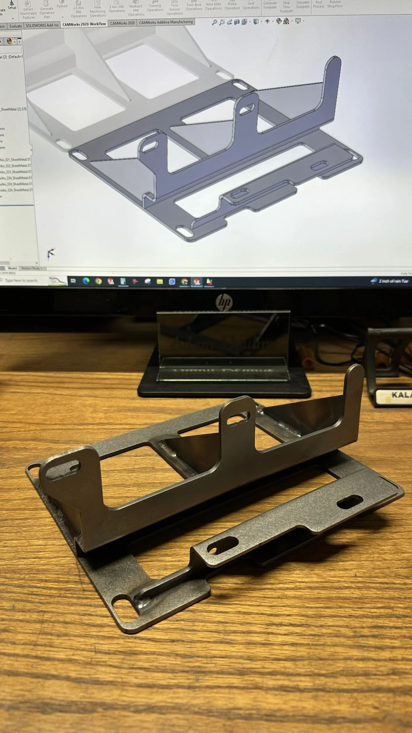 A computer-aided design (CAD) drawing of a metal component is displayed on a monitor, with a physical metal piece resembling the design placed on a wooden desk in front of it. The CAD image shows a detailed 3D model of a bracket or mount with cutouts and bends.