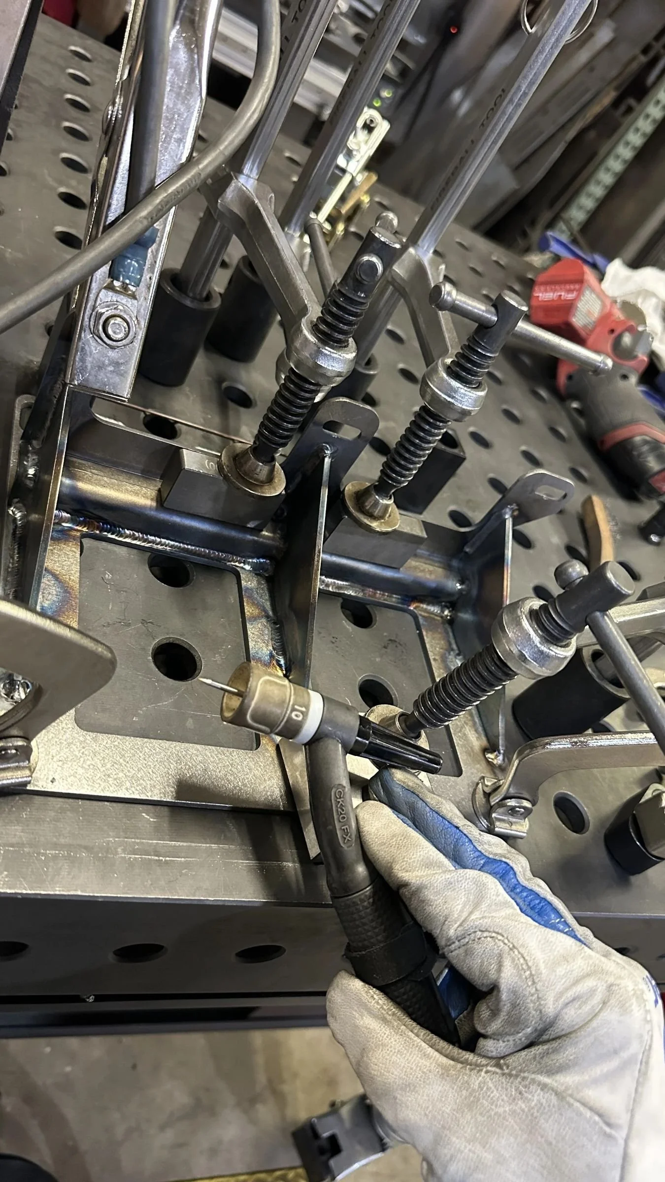 A person wearing white gloves is using a welding tool on a metal structure with various screws, rods, and mechanical components in a workshop.