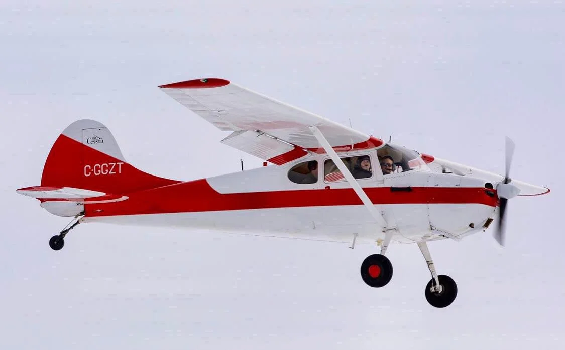 A small, white and red propeller airplane flying in the sky with two pilots visible inside.