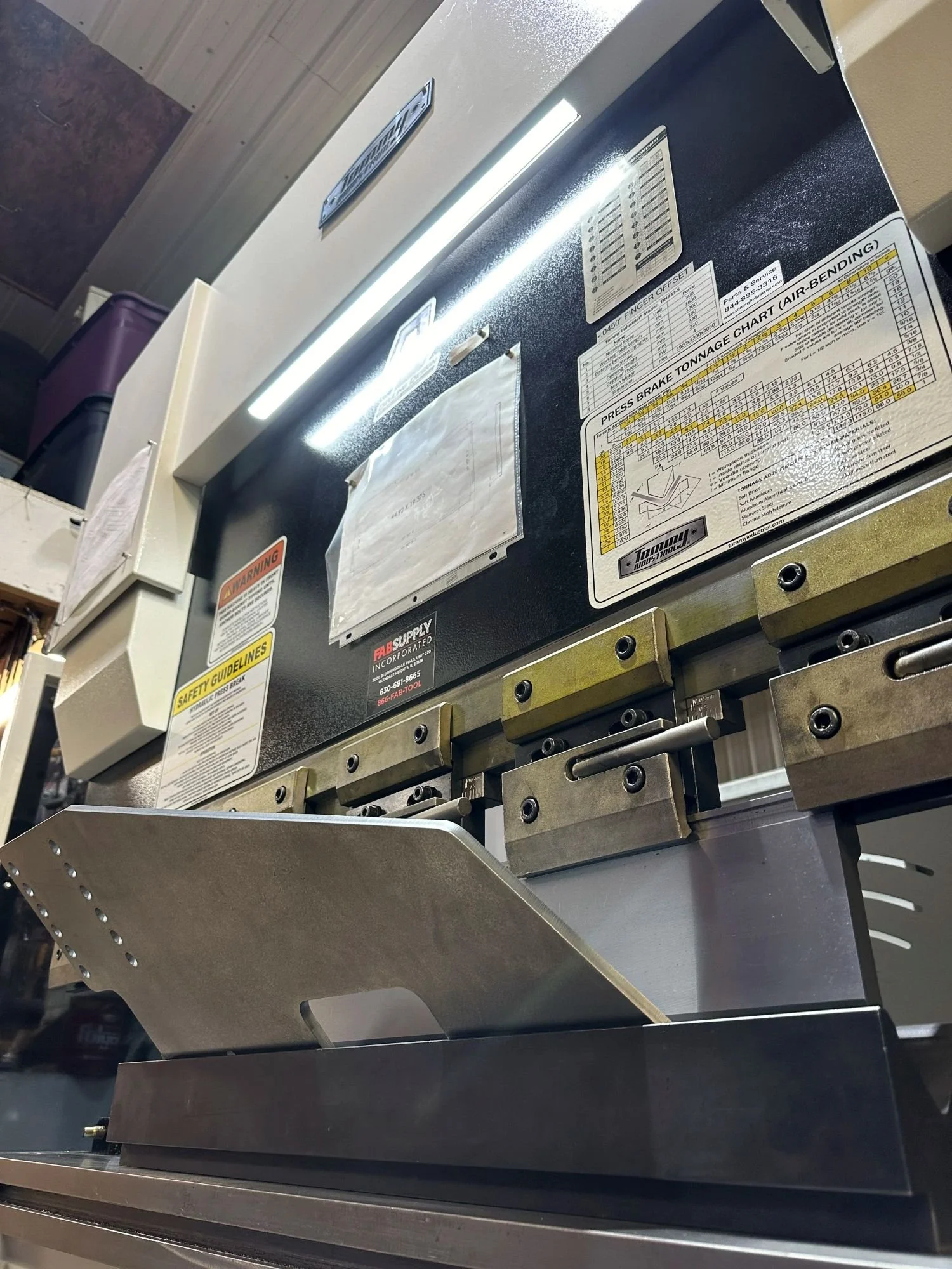 Close-up of an industrial metal press brake bending machine with safety and operation labels, bending a metal component.