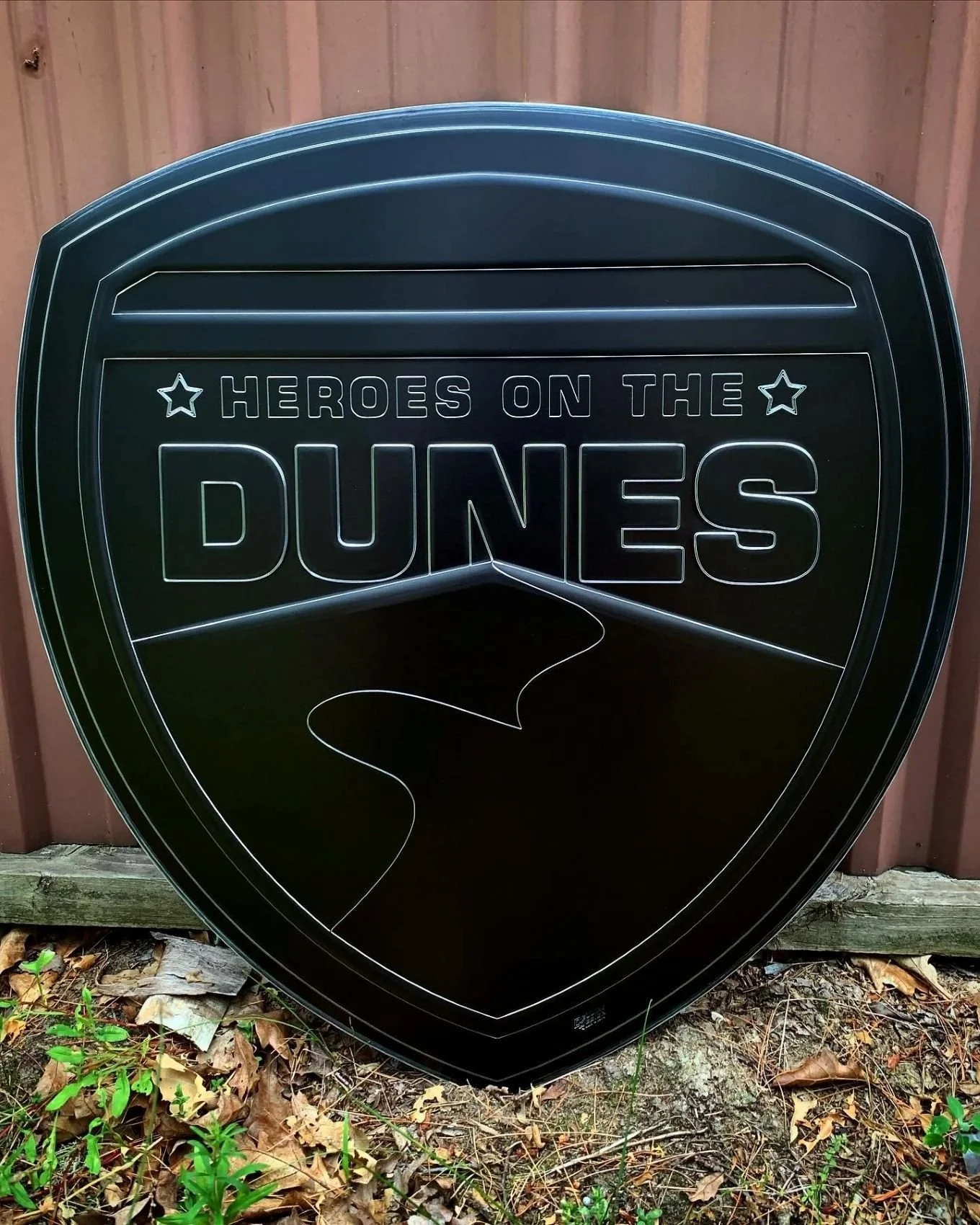 Black metal sign shaped like a superhero emblem with the text 'Heroes on the Dunes' propped outside.