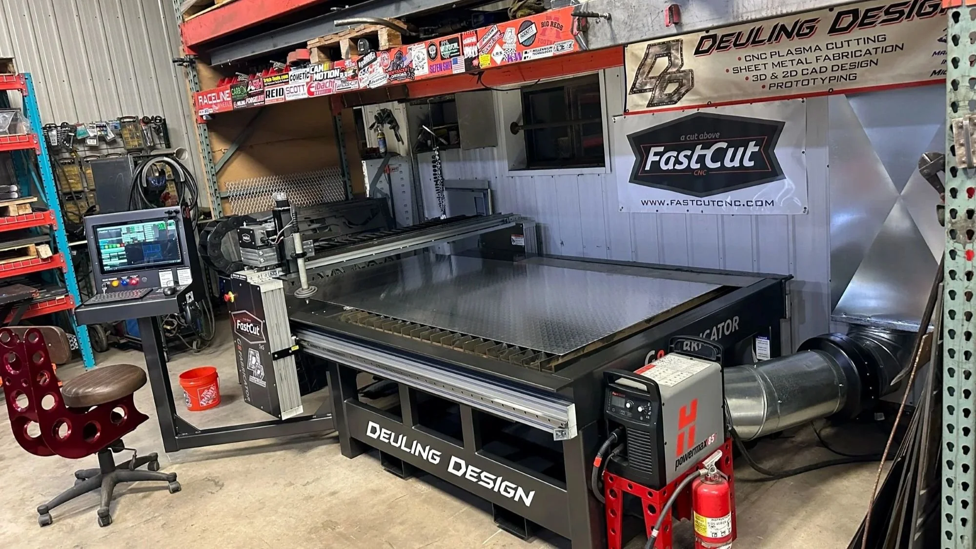 A CNC plasma cutting machine in a workshop with a computer control panel, surrounded by shelves with tools and stickers on the wall.