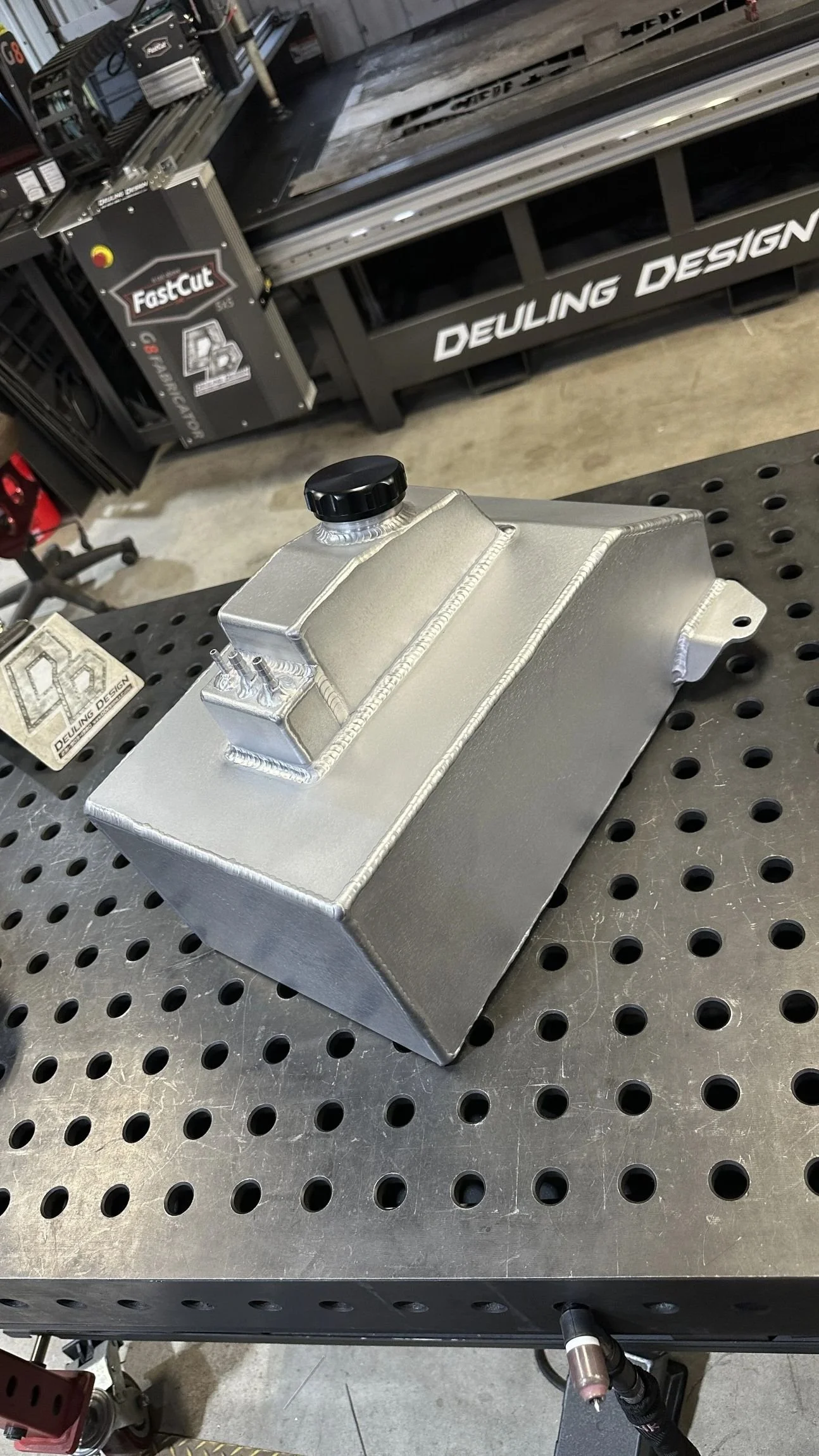 Custom aluminum tank with precision welds, a threaded filler cap, and mounting tabs, displayed on a metal welding table.