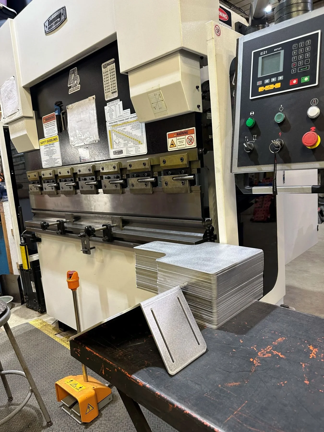 Industrial metal press brake bending machine with control panel, metal sheets stacked on a table, and tools nearby in a workshop setting.