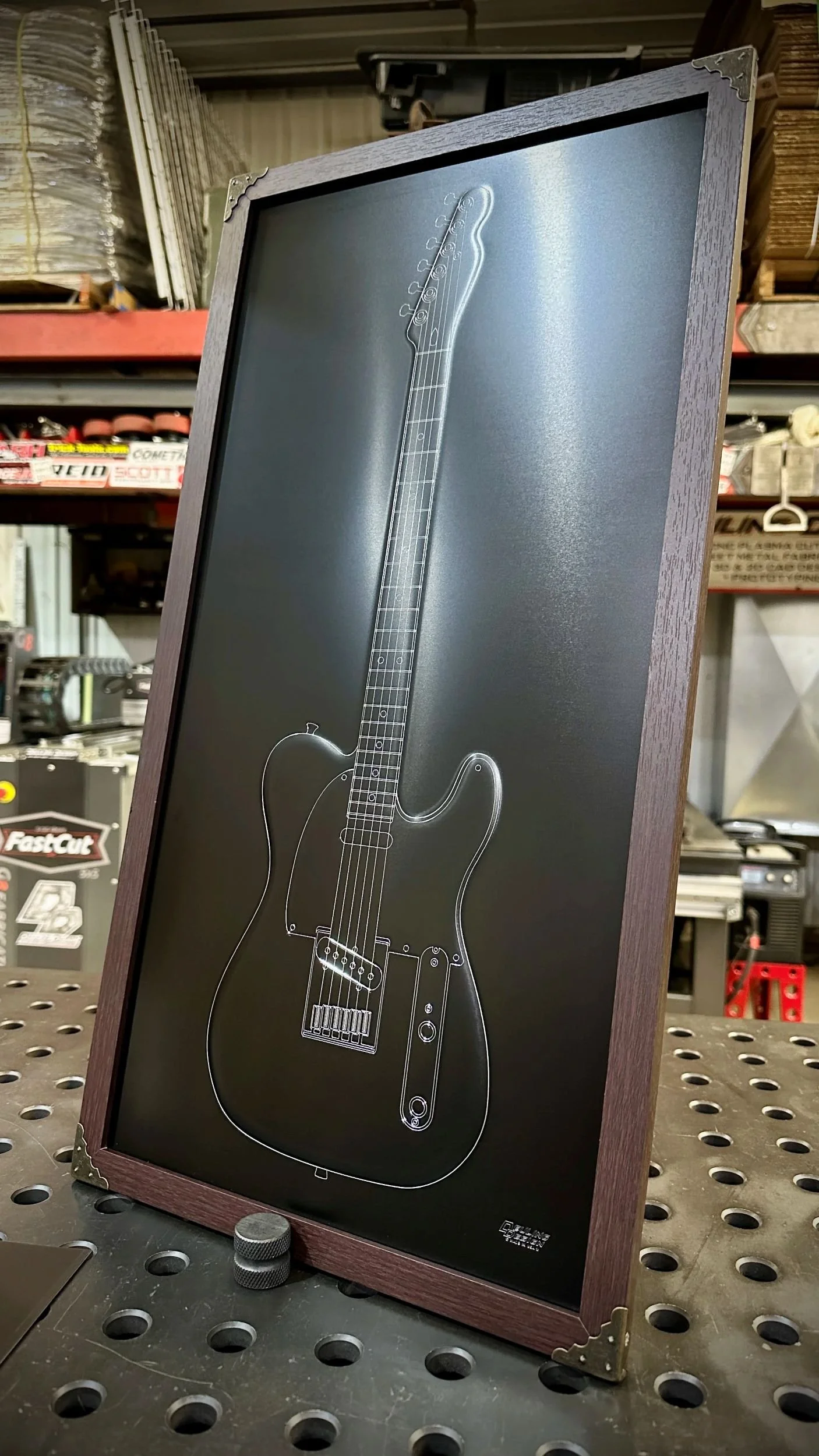 Framed artwork of a beadroll electric guitar outline on black metal, leaning against a metal surface in a workshop.
