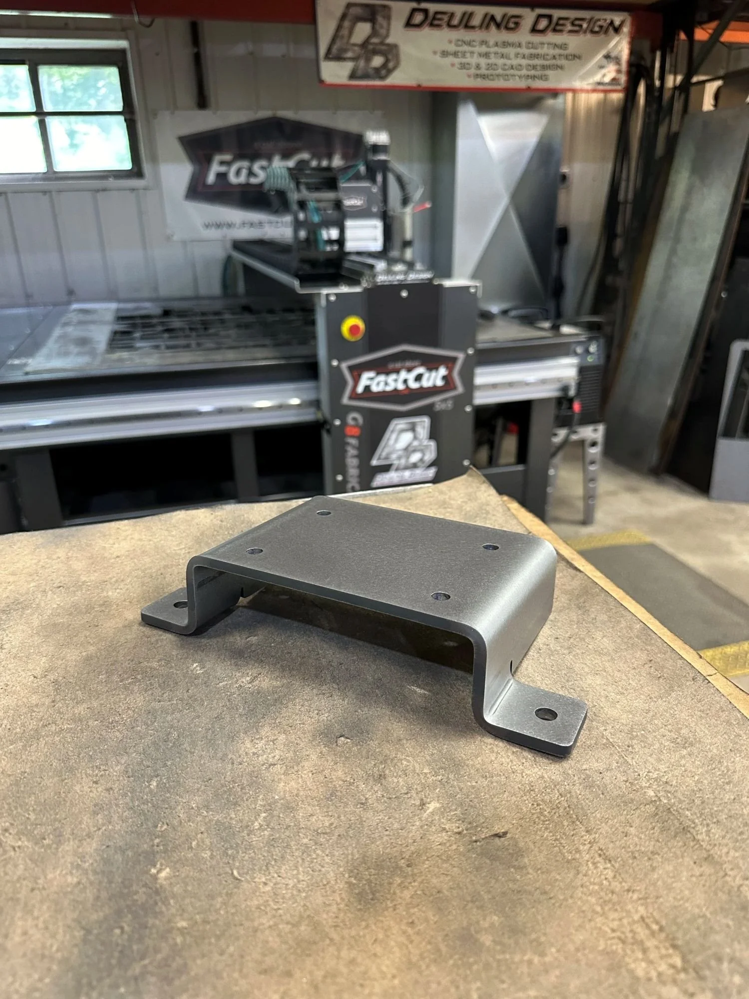 A metal bracket resting on a workbench in a workshop. In the background, there are signs for Deuling Design and FastCut CNC machine.