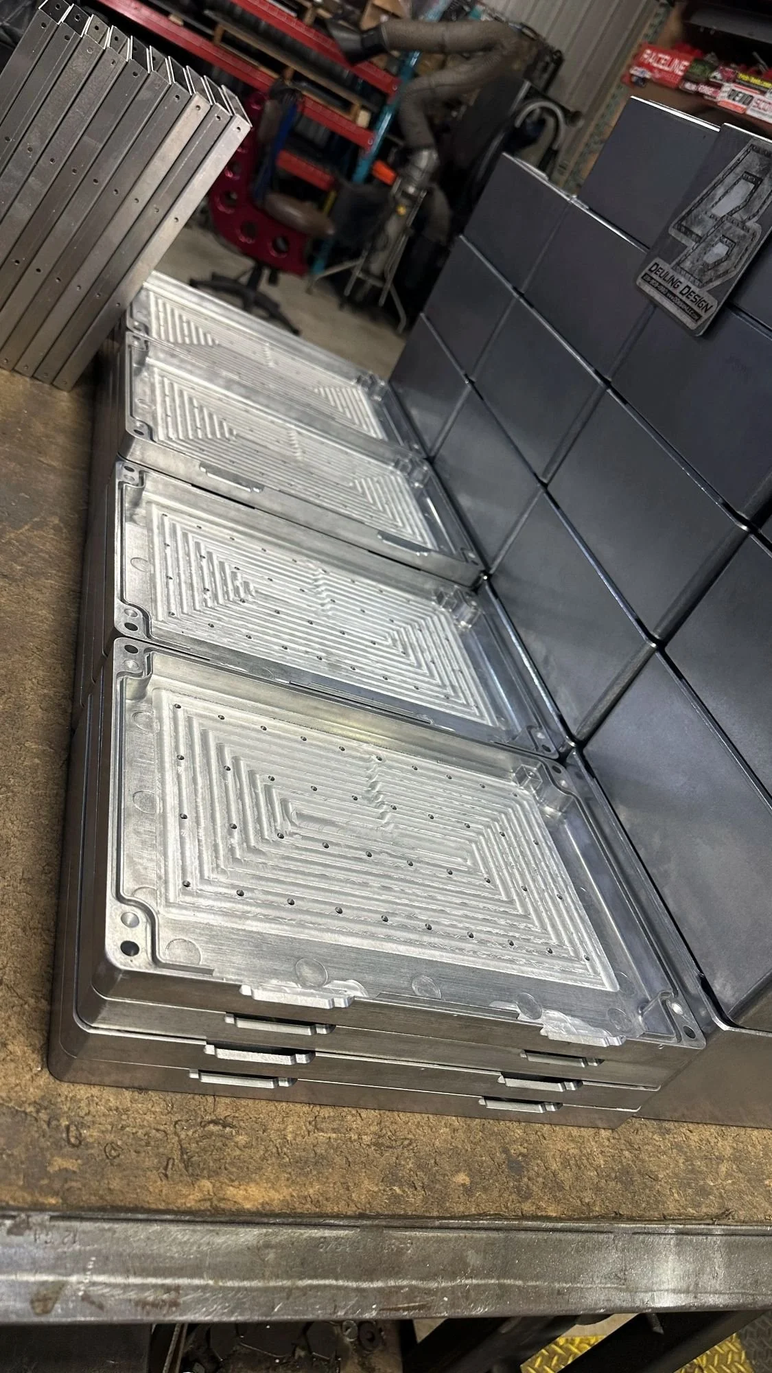 Stacked metallic rectangular parts with detailed patterns, possibly machine parts or electronic enclosures, on a workbench in a workshop with tools and equipment in the background.