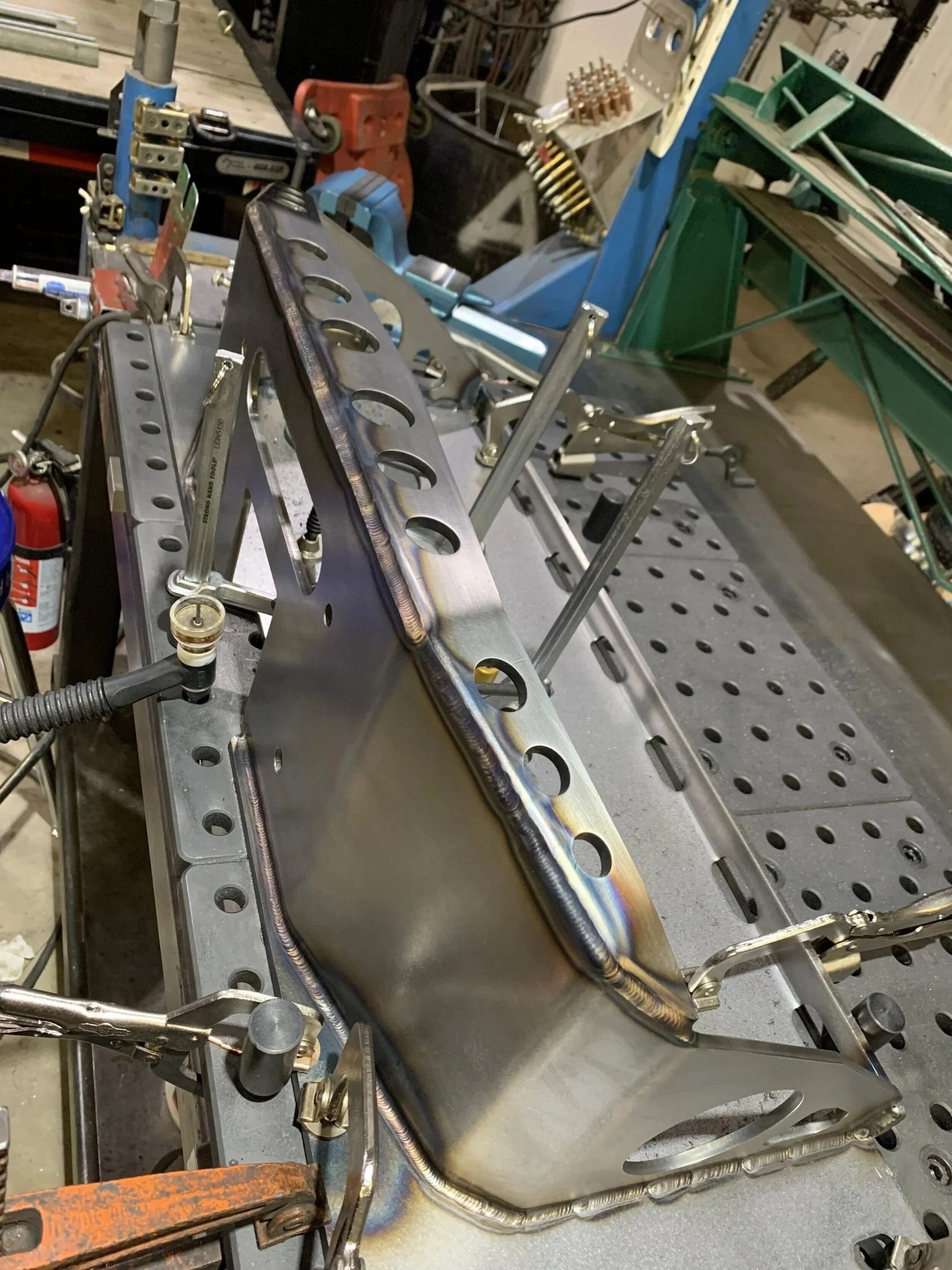 A metal workbench with a welded metal plate and various metal tools in a workshop. The background shows tools, containers, and other workshop equipment.