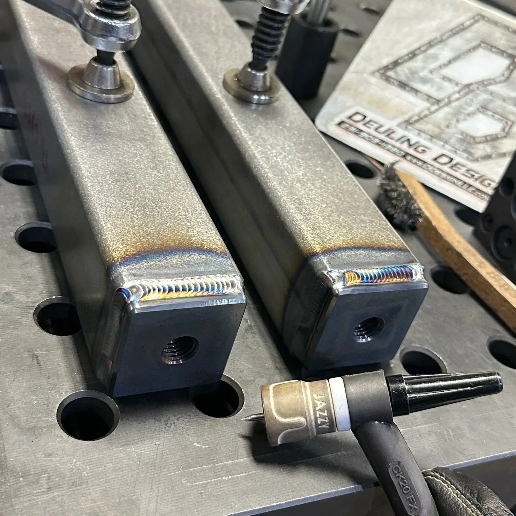 Close-up of two metal component ends welded, including a welding tool and Deuling Design logo in background.
