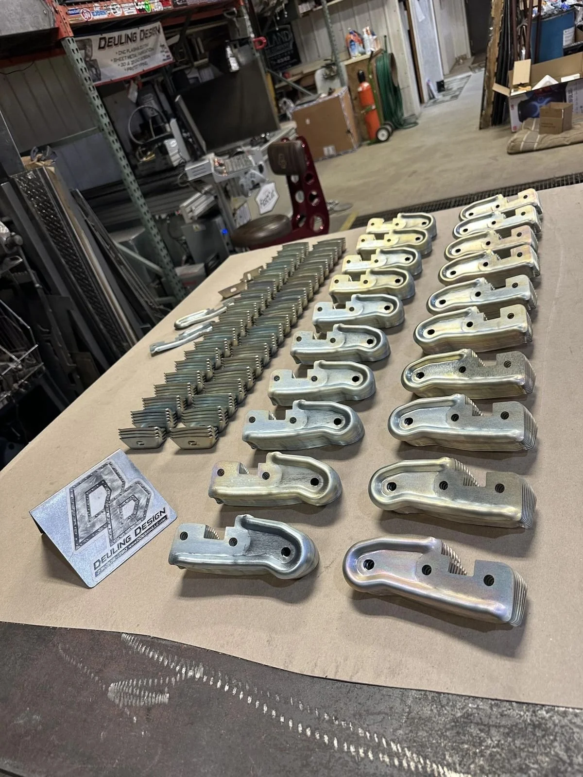 Collection of metal parts arranged on a worktable in a workshop, with shelving, tools, and supplies in the background.