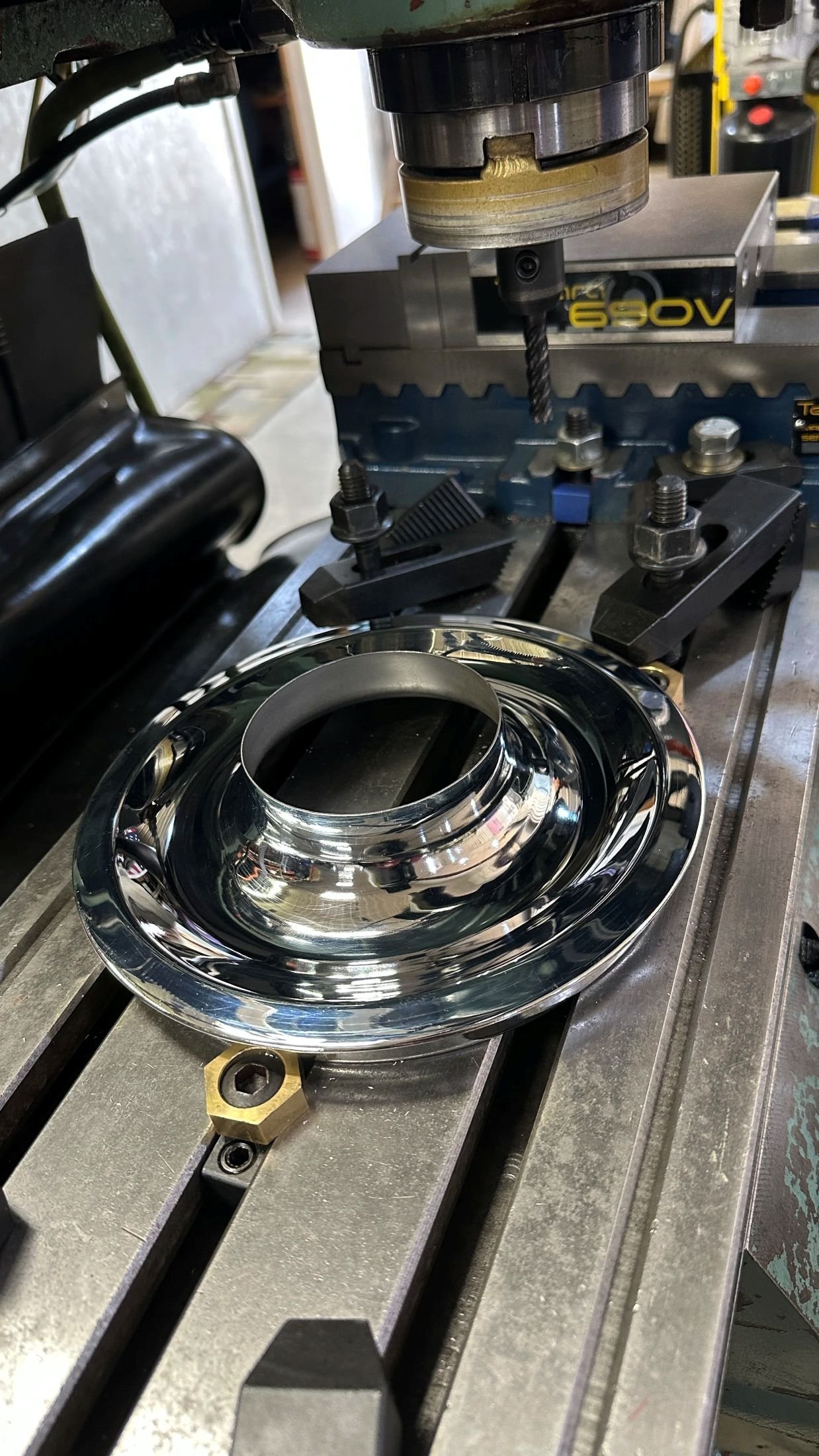 A metal machining setup with a drill bit mounted on a machine, positioned above a shiny, reflective metal ring resting on a metallic work surface.