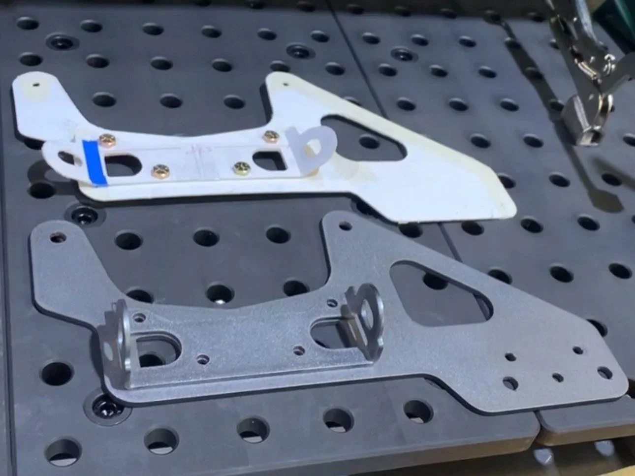 Metal brackets and mounting plates on a perforated workbench, with a tool partially visible in the upper right corner.