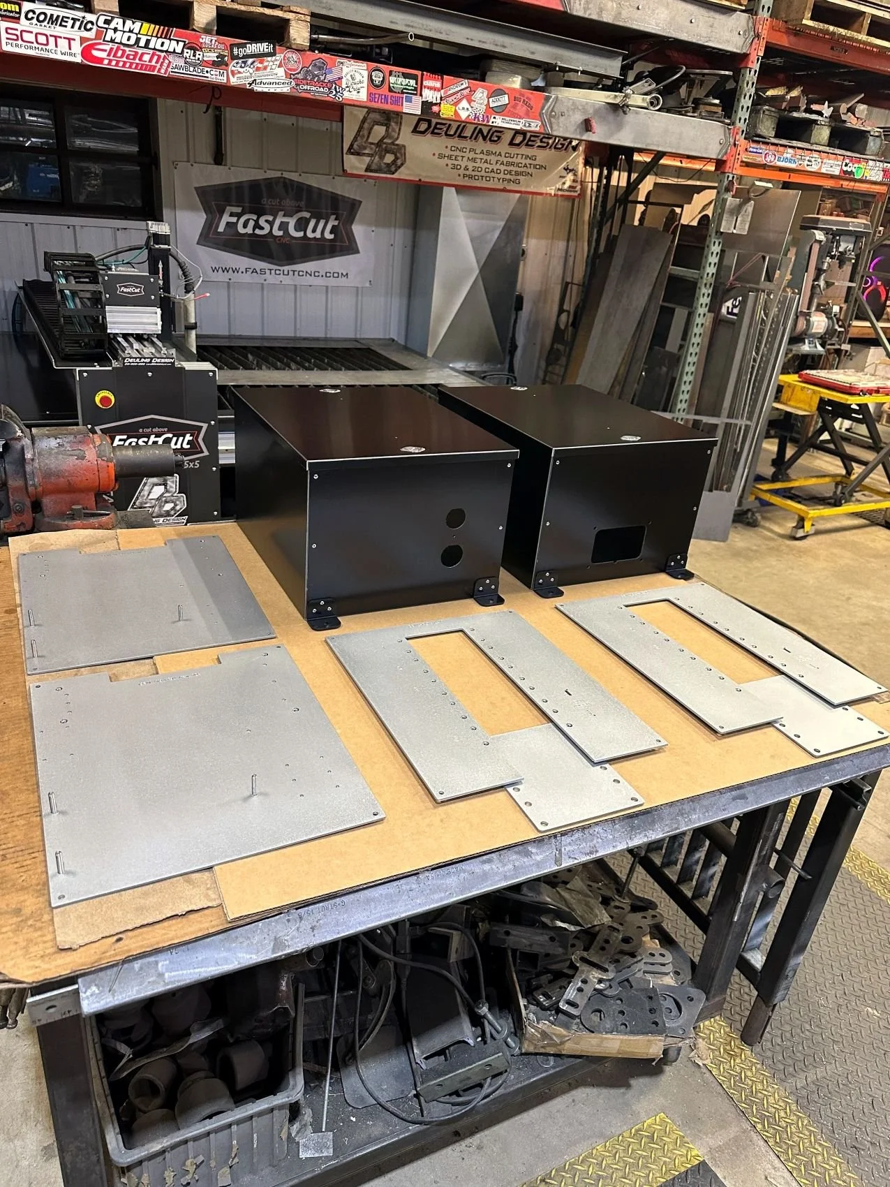 Workshop workspace with metal sheets, tools, and equipment on a workbench, shelves with stickers and signs, metal parts, and a yellow rubber mat on the floor.
