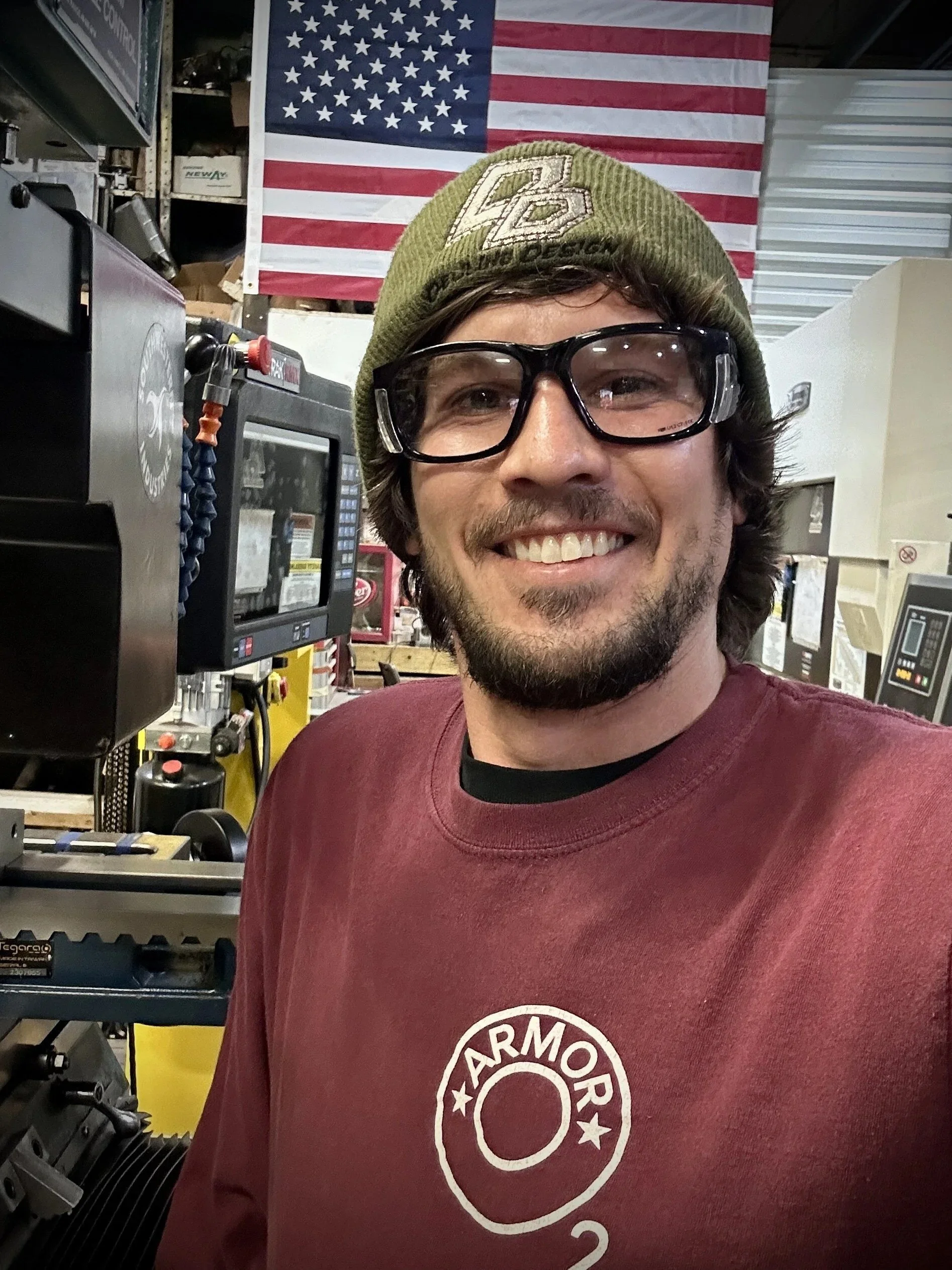 A man wearing a green beanie hat, black glasses, and a maroon shirt with a circular logo, smiling in a workshop with American flag in the background.