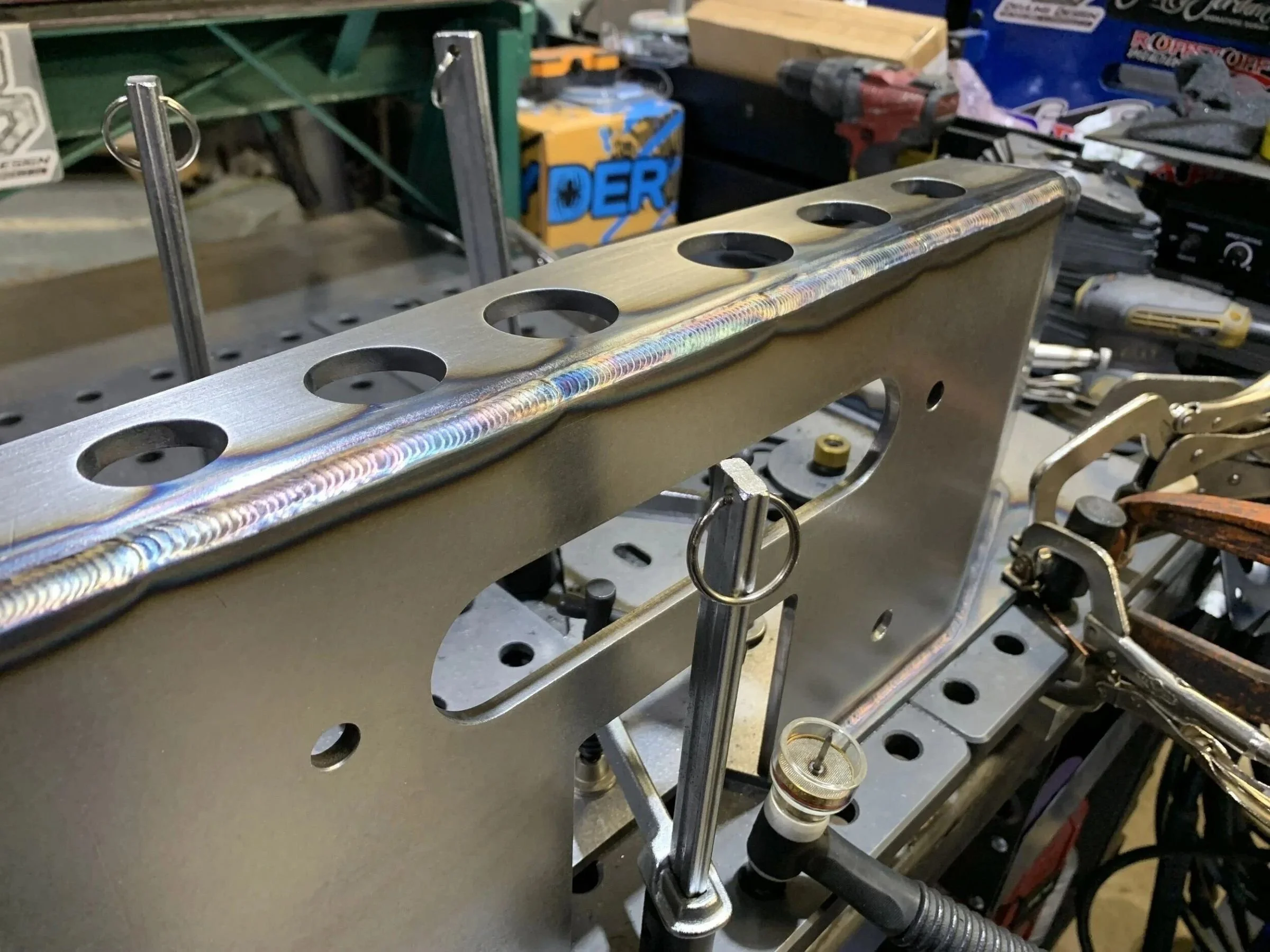 Close-up of a steel fabrication assembly clamped in a welding fixture, featuring a clean weld bead along the top edge.