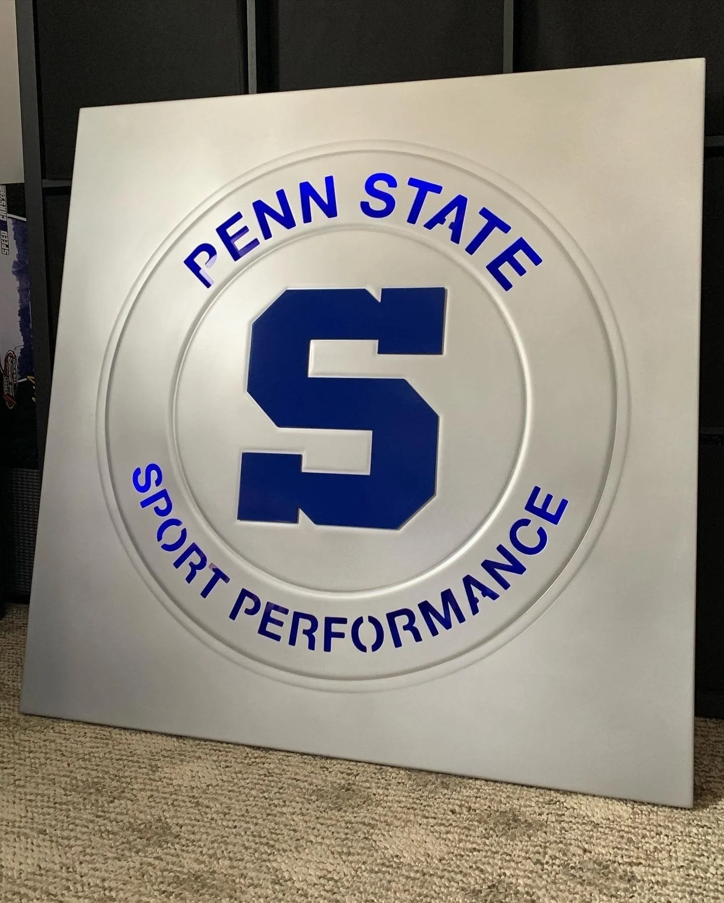 A metal sign with 'PENN STATE' and 'SPORT PERFORMANCE' in a circular design with a large 'S' in center. Verbiage is backlit with blue.