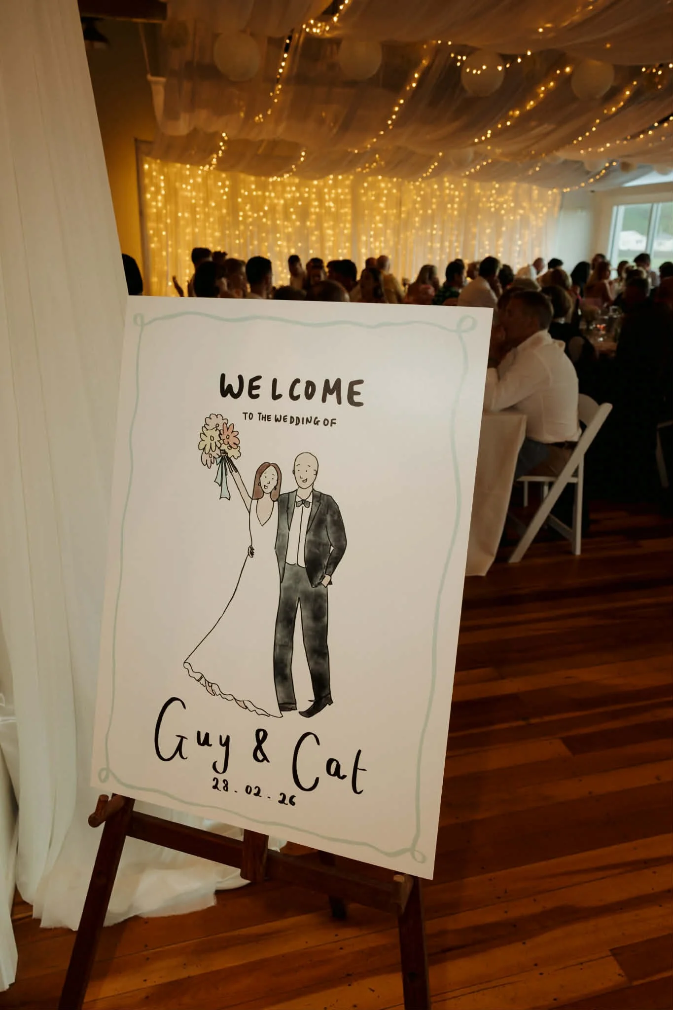 A wedding reception with a sign in the foreground that features a drawing of a bride and groom. The bride is holding a bouquet. The sign reads 'WELCOME TO THE WEDDING OF Guy & Cat 28.02.26' with a softly lit background of hanging lights and paper lanterns, and guests seated at tables in the background.