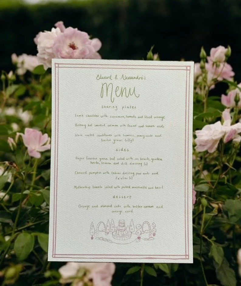 A wedding menu card with handwritten text, placed among pink and white flowers and green foliage.
