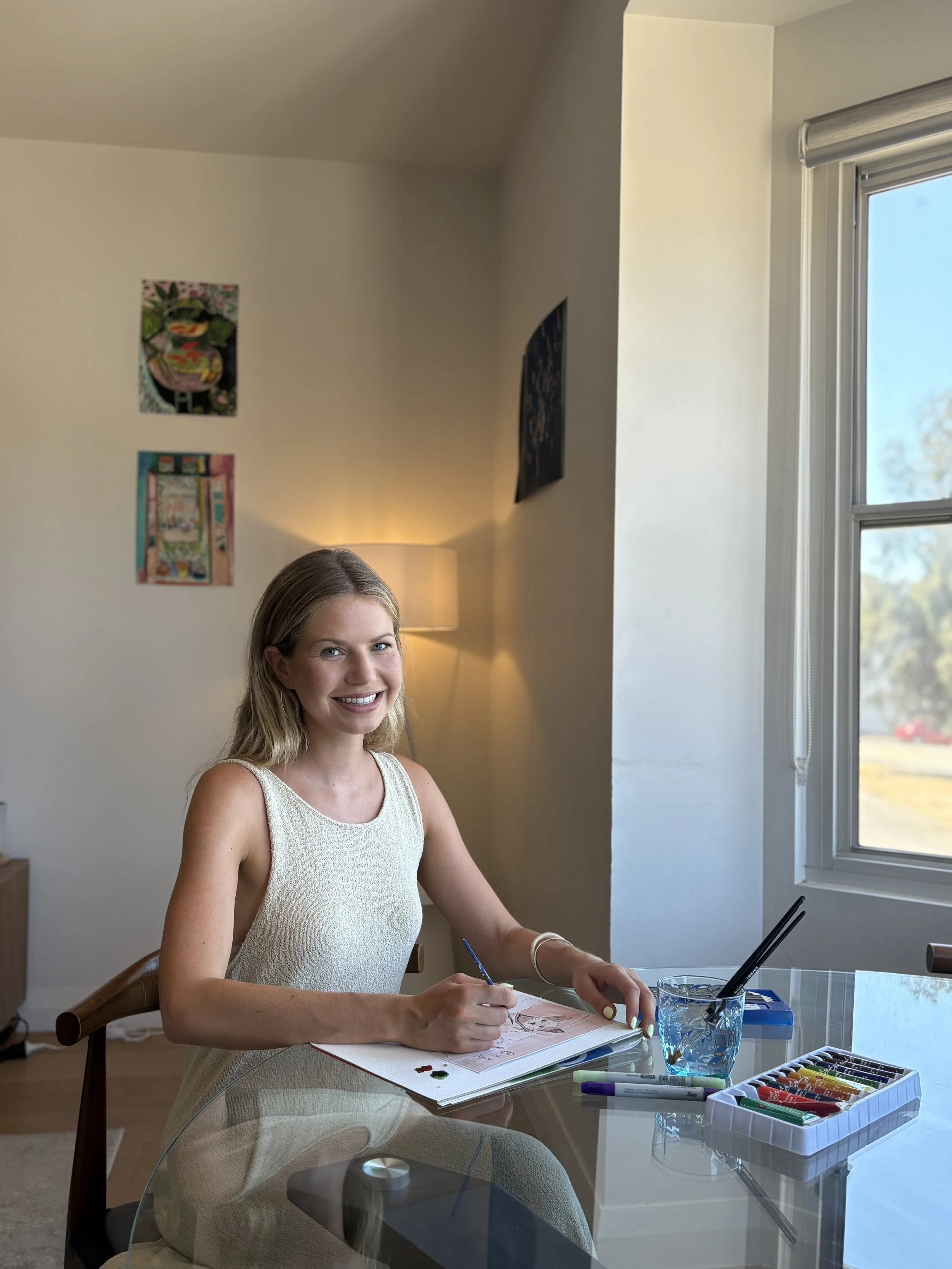 Young woman with blonde hair sitting at a glass table, smiling and sketching in a notebook. Art supplies such as markers and colored pencils are on the table. Sunlight streams through a window on the right side, illuminating the room decorated with pictures on the wall.
