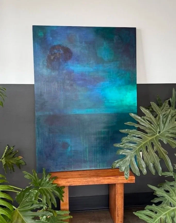 Abstract painting with various shades of blue and teal, mounted on a wall above a wooden stand, with green leafy plants in the foreground.