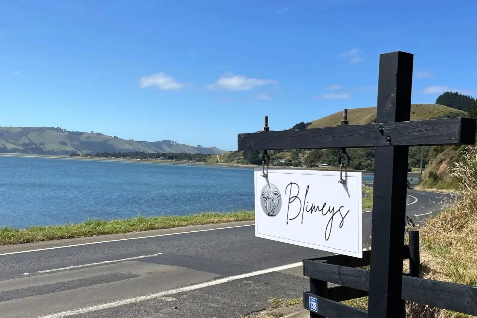 A lakeside road scene with a black wooden fence and a sign that says 'Blumys' hanging from it, overlooking a calm lake with rolling green hills in the background under a clear blue sky.