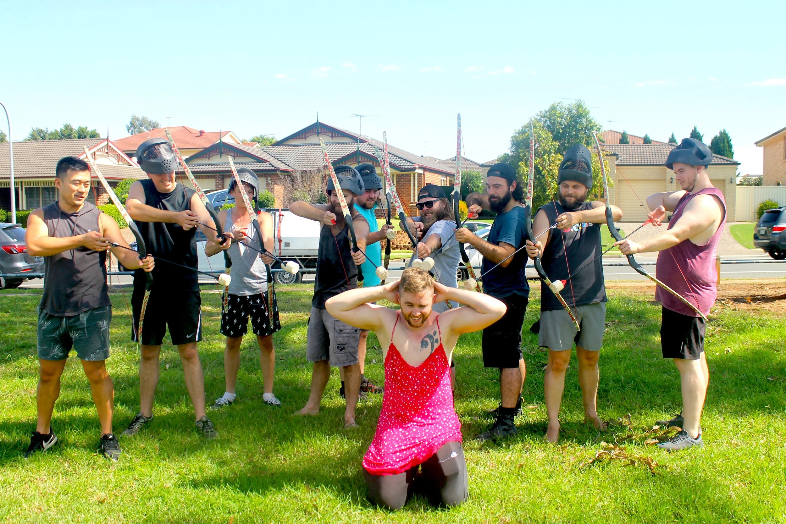 Group of ten young men playing archery tag outdoors on a grassy field, with houses and trees in the background, one man, dressed in a red dress, kneeling in front holding his head and smiling, others wearing helmets and casual clothes aiming bows.