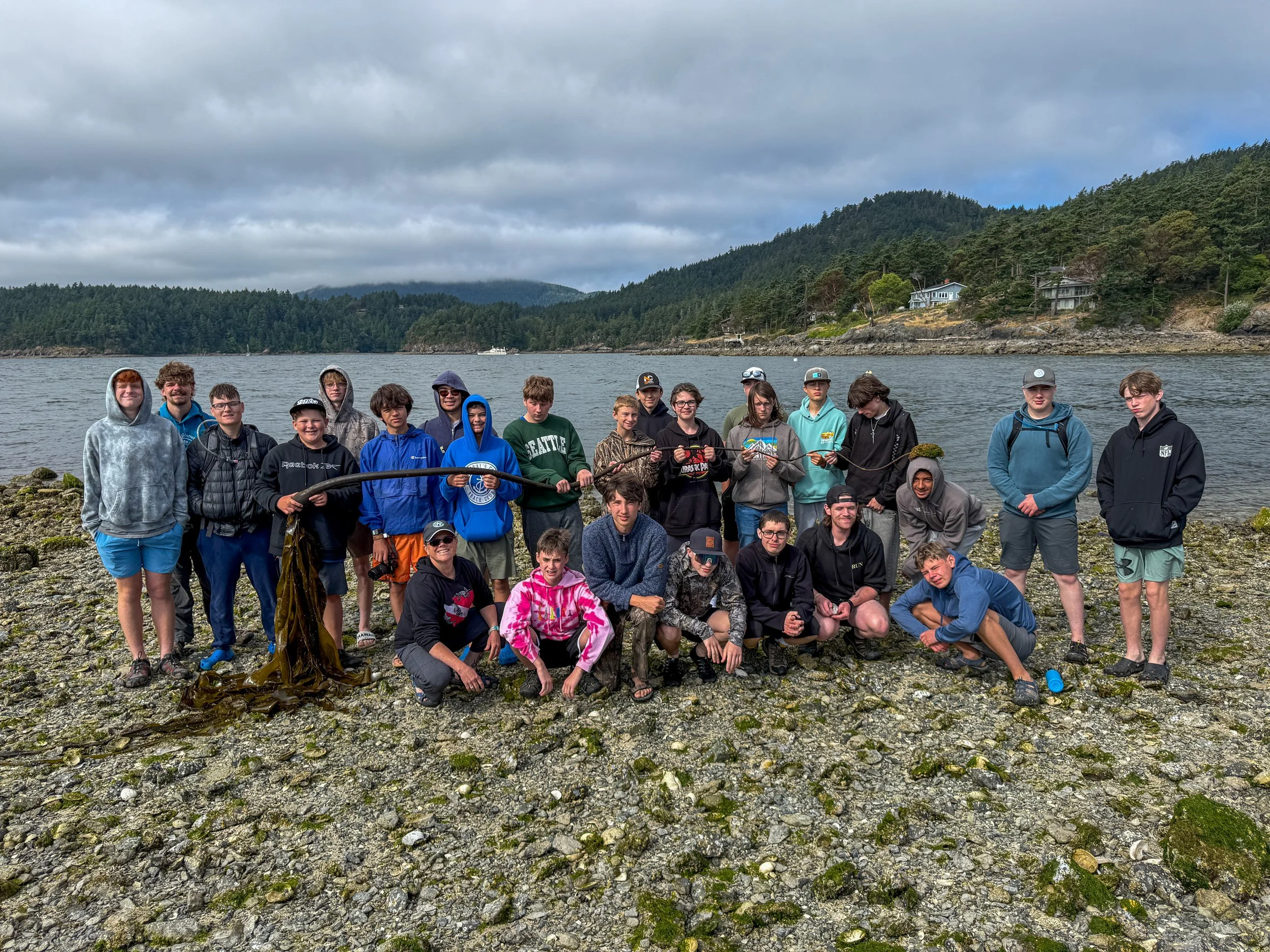 Group of young people posing on rocky shoreline near water with forested hills in the background.