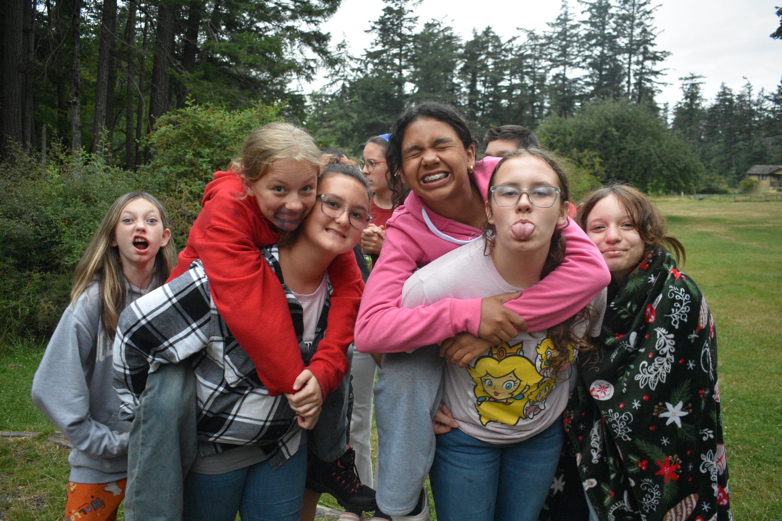 A group of young girls outdoors making funny faces and showing playful expressions, with a forest background.