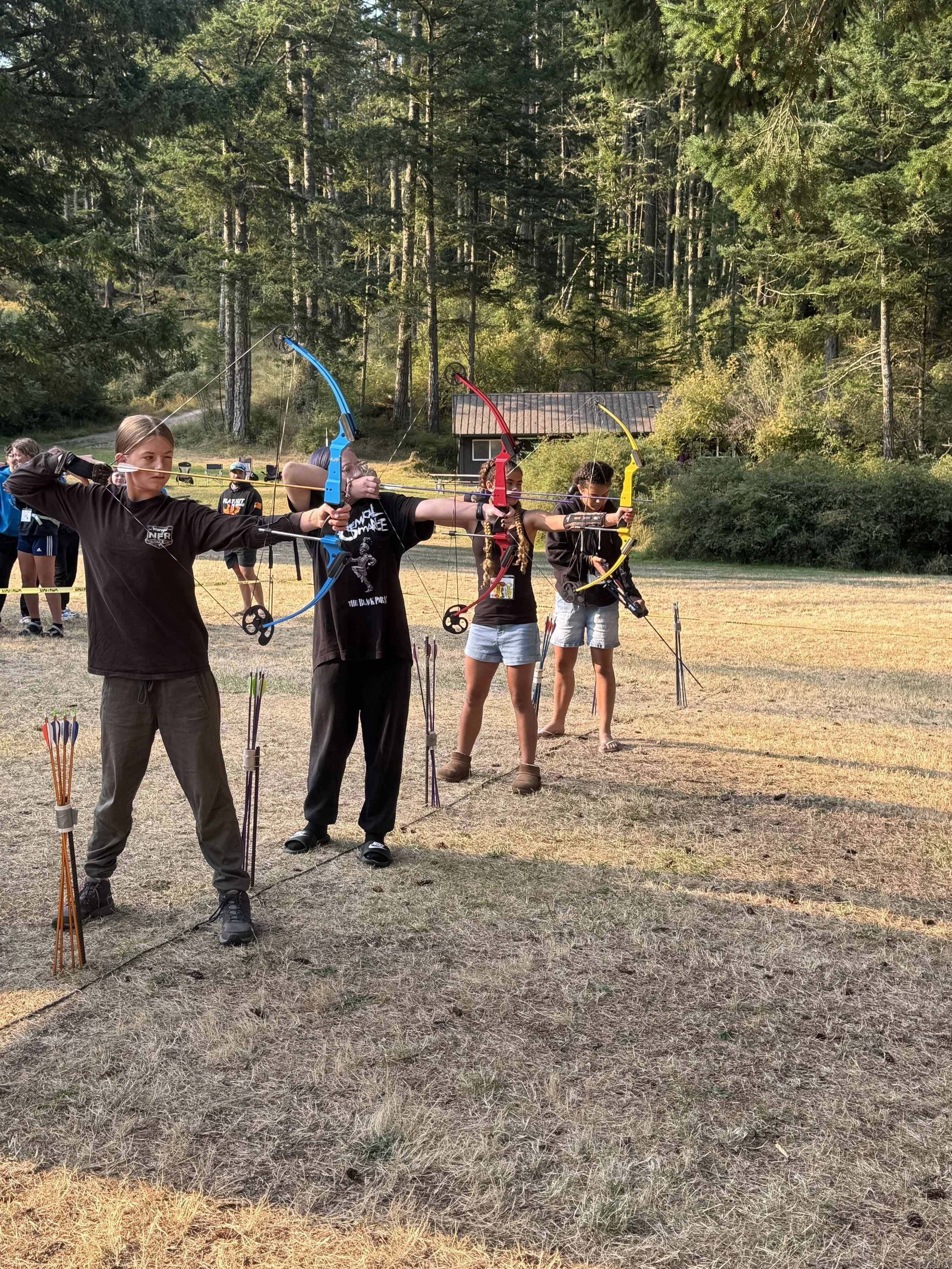 Group of people practicing archery outdoors in a grassy field with trees in the background.