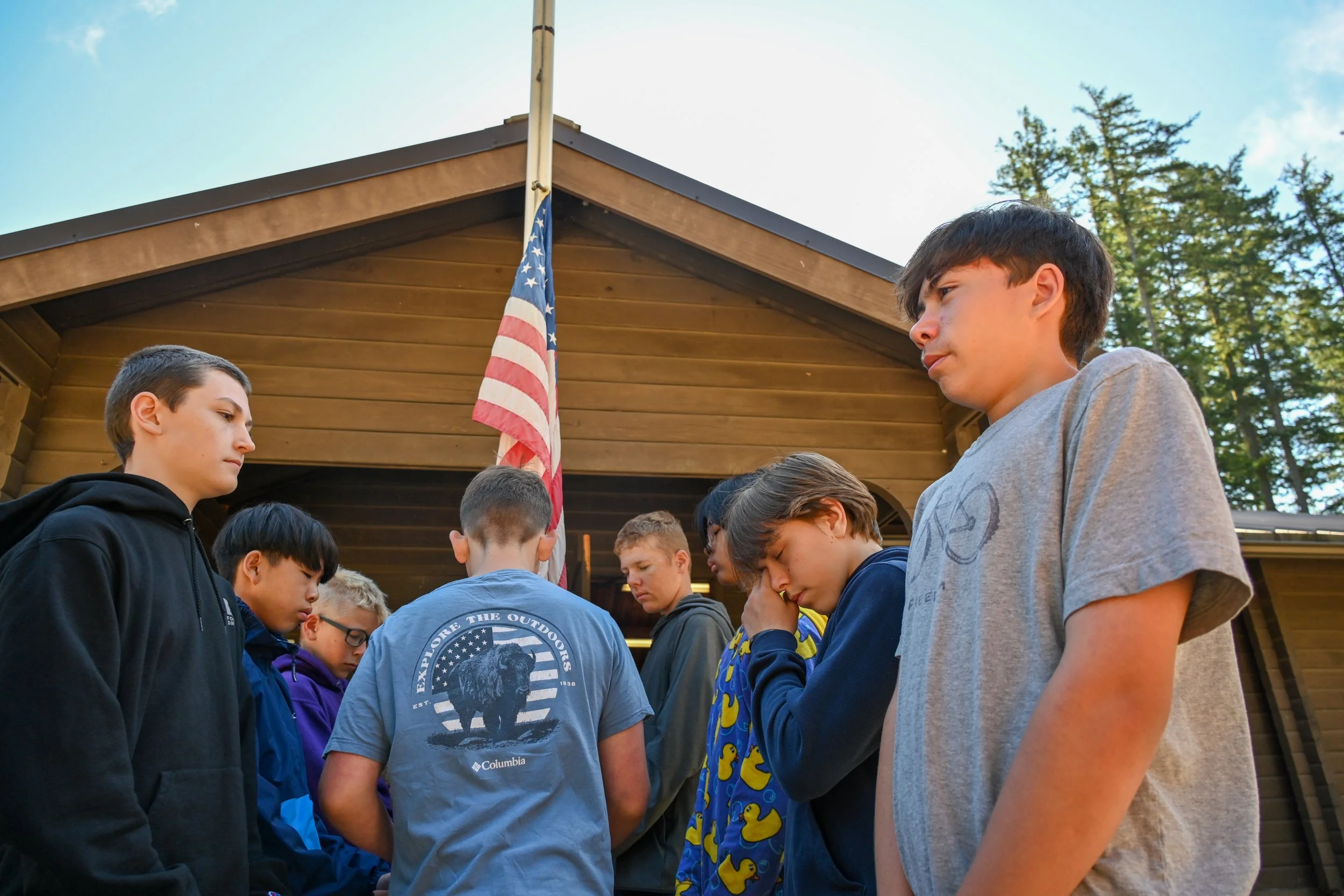 A group of young boys standing outside in front of a wooden building with an American flag, some seem to be praying or reflecting.