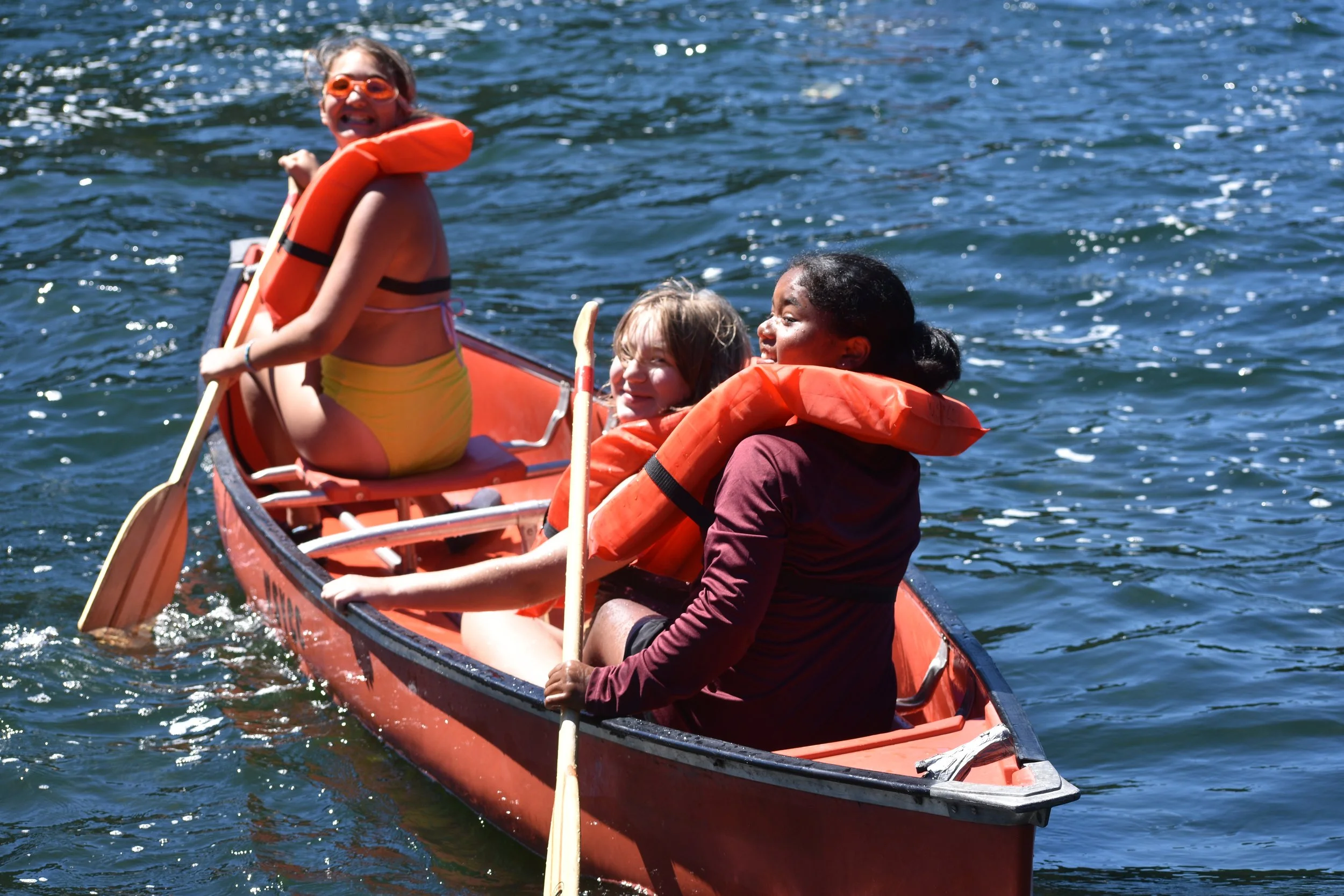 Three people in a red canoe on the water, wearing orange life jackets. One person at the front smiling, and two others in the middle and back paddling.