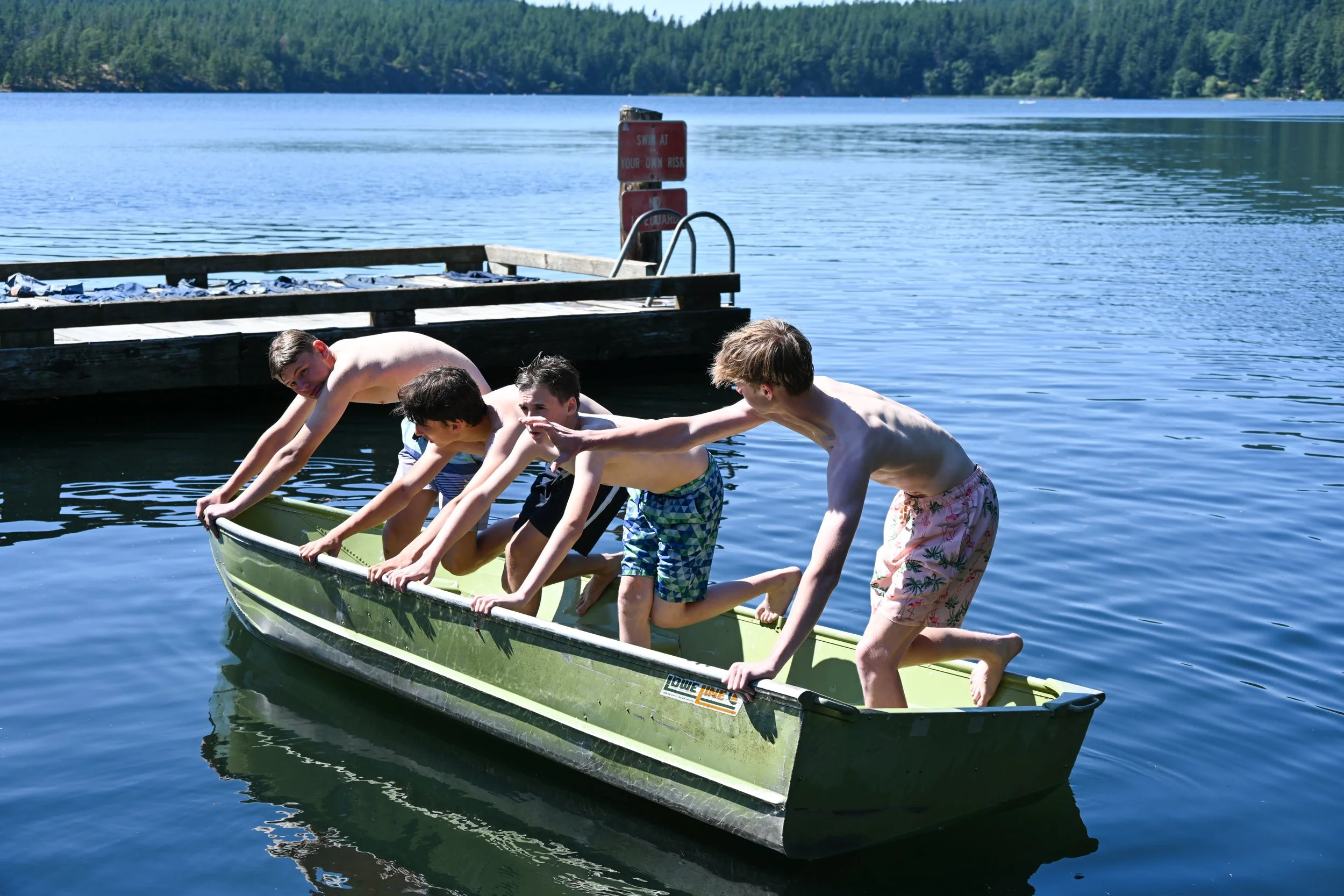 Four boys are pushing a small green boat into a lake from a dock, surrounded by trees under a clear blue sky.
